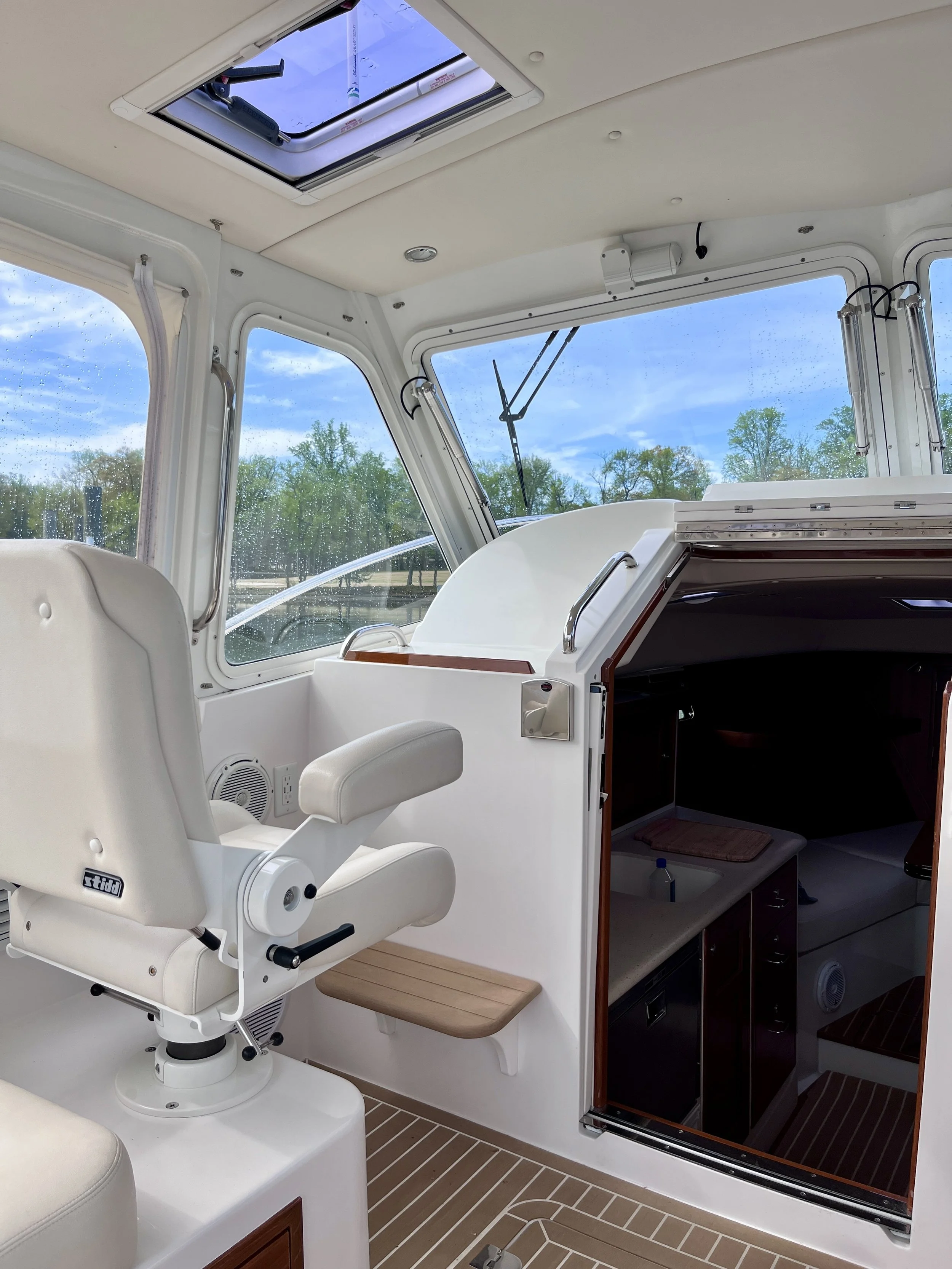 Inside view of a boat, showing a white reclining captain's seat, open ceiling hatch with a flashlight, large windows with rain droplets, and a sliding door leading to the cabin with wooden flooring.