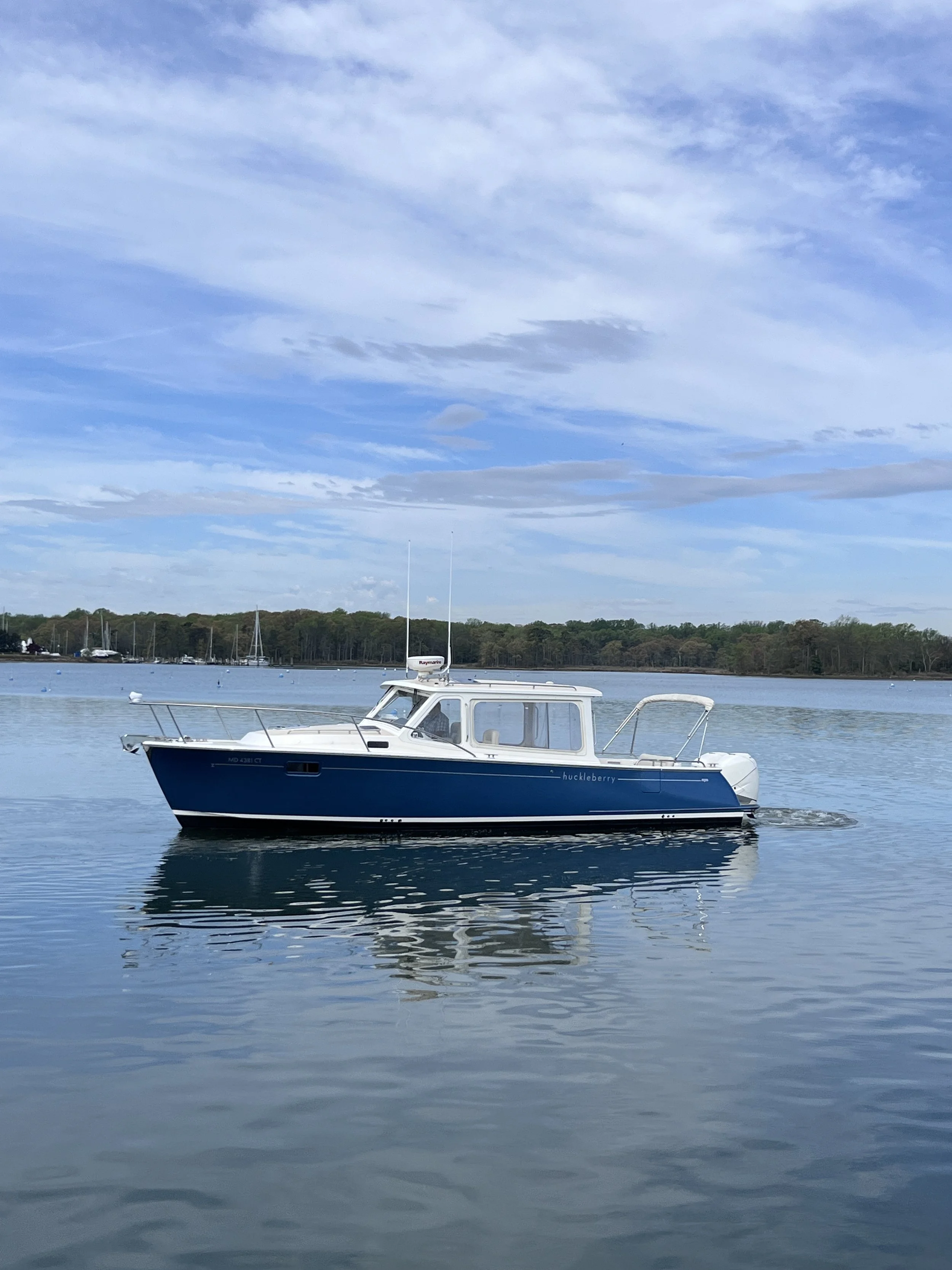 A boat on calm water with a distant shoreline lined with trees and a partly cloudy sky overhead.