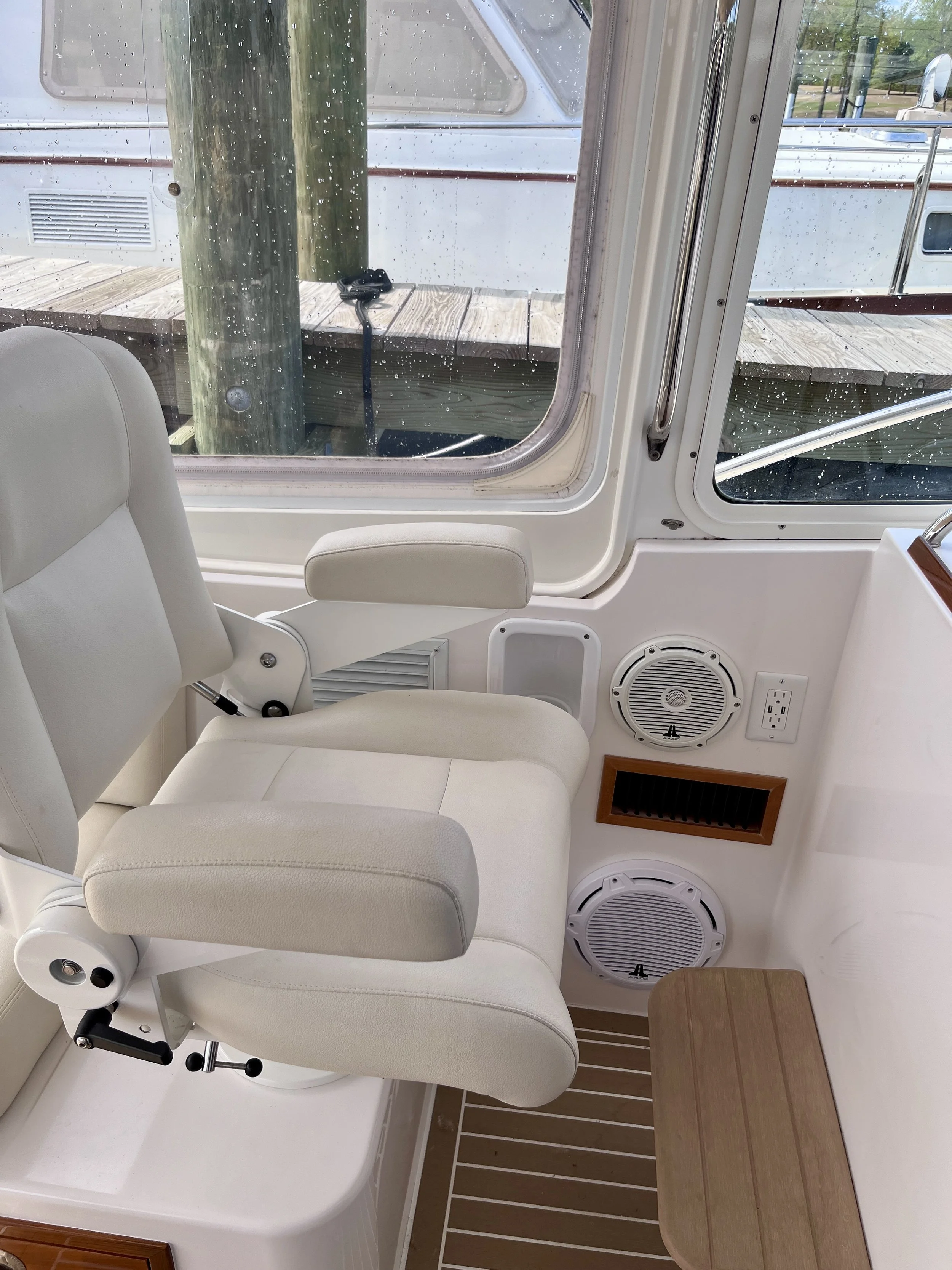 Inside a boat, view of two white captain chairs with armrests, a small wooden table, and a window with rain droplets on the glass, showing a dock and boat outside.