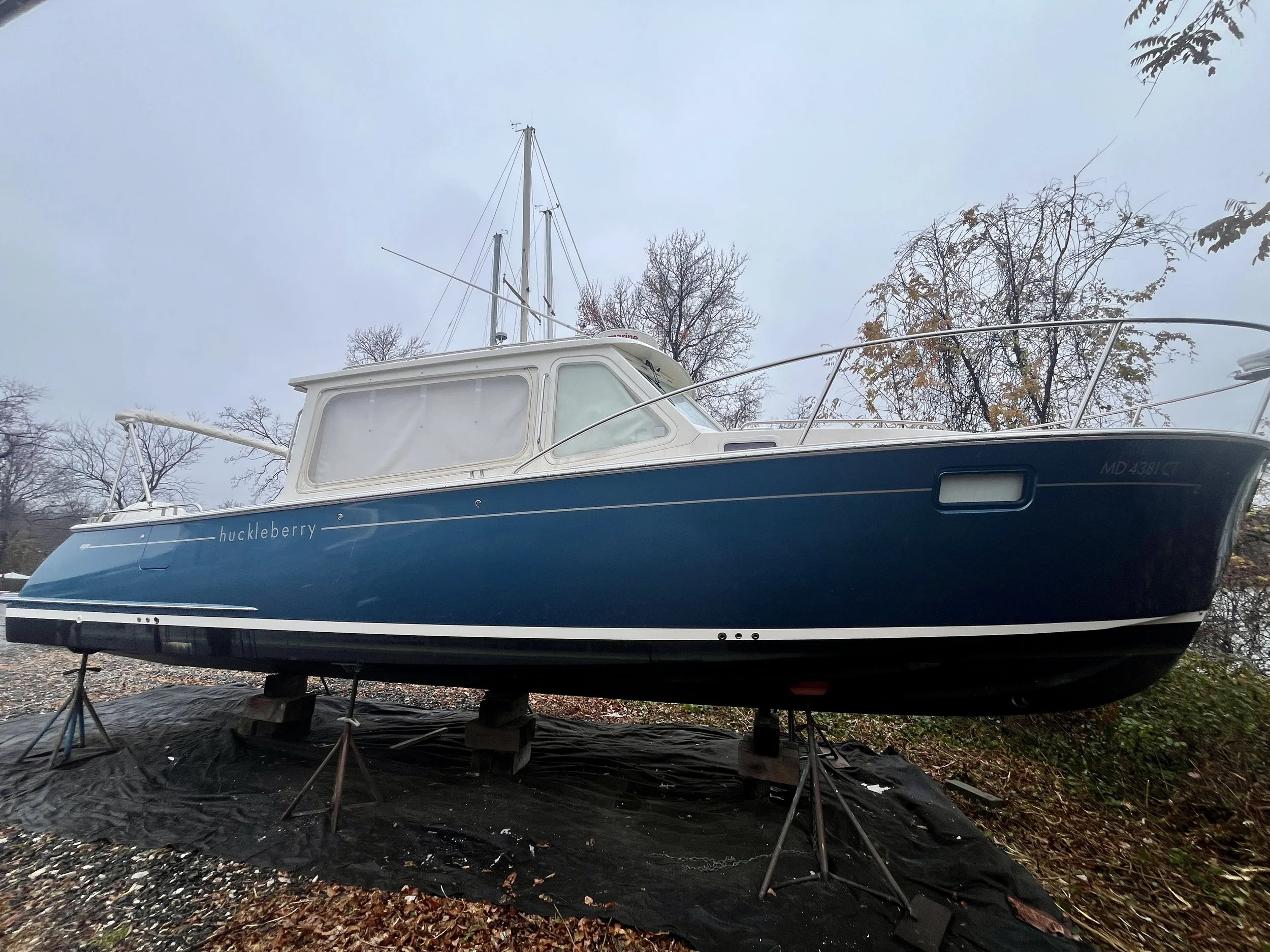 A boat named 'huckleberry' is on land, supported by wooden blocks, with trees and an overcast sky in the background.