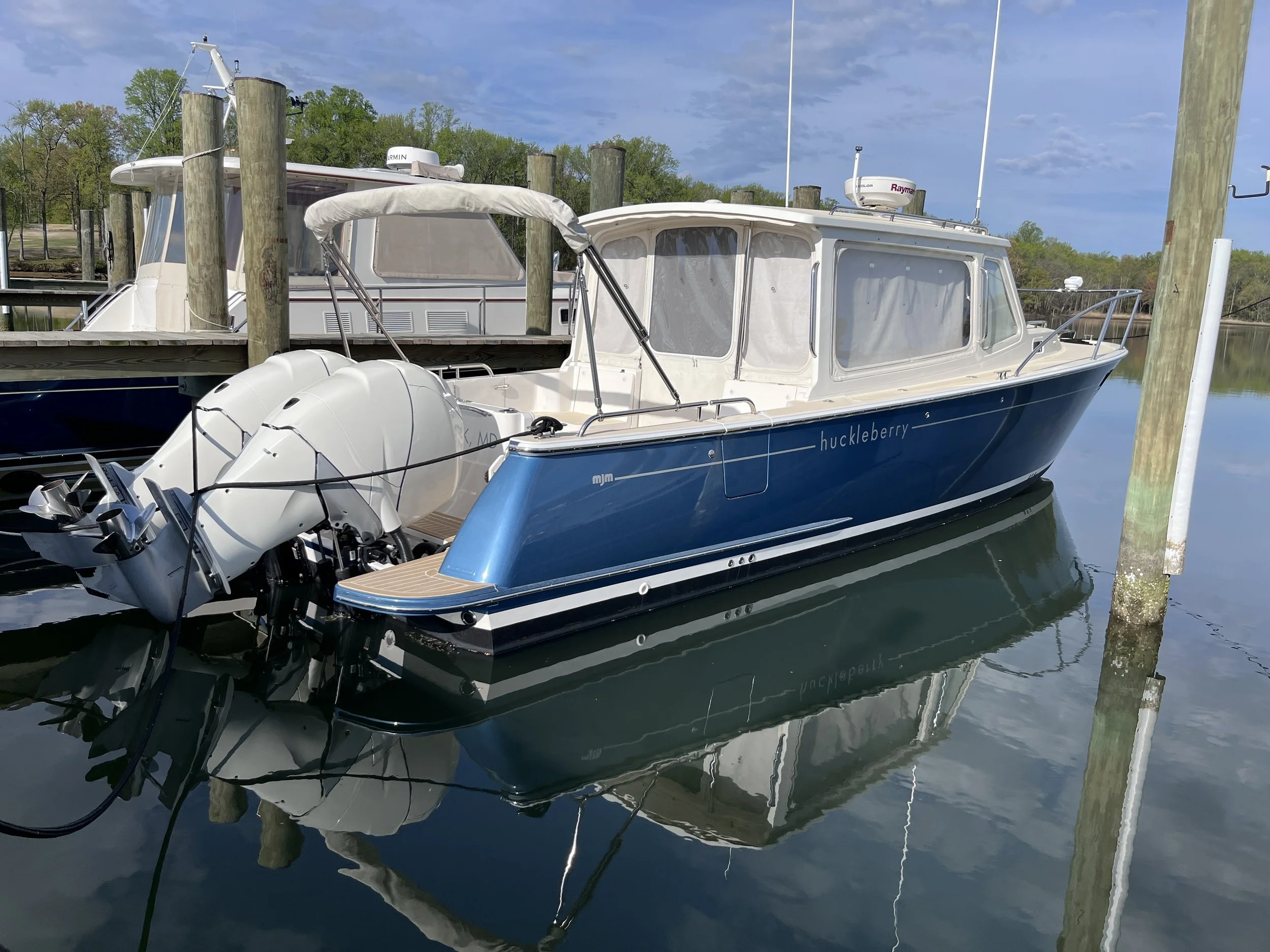 A blue and white boat named 'huckleberry' docked at a marina with wooden pilings, calm water, and green trees in the background under a partly cloudy sky.
