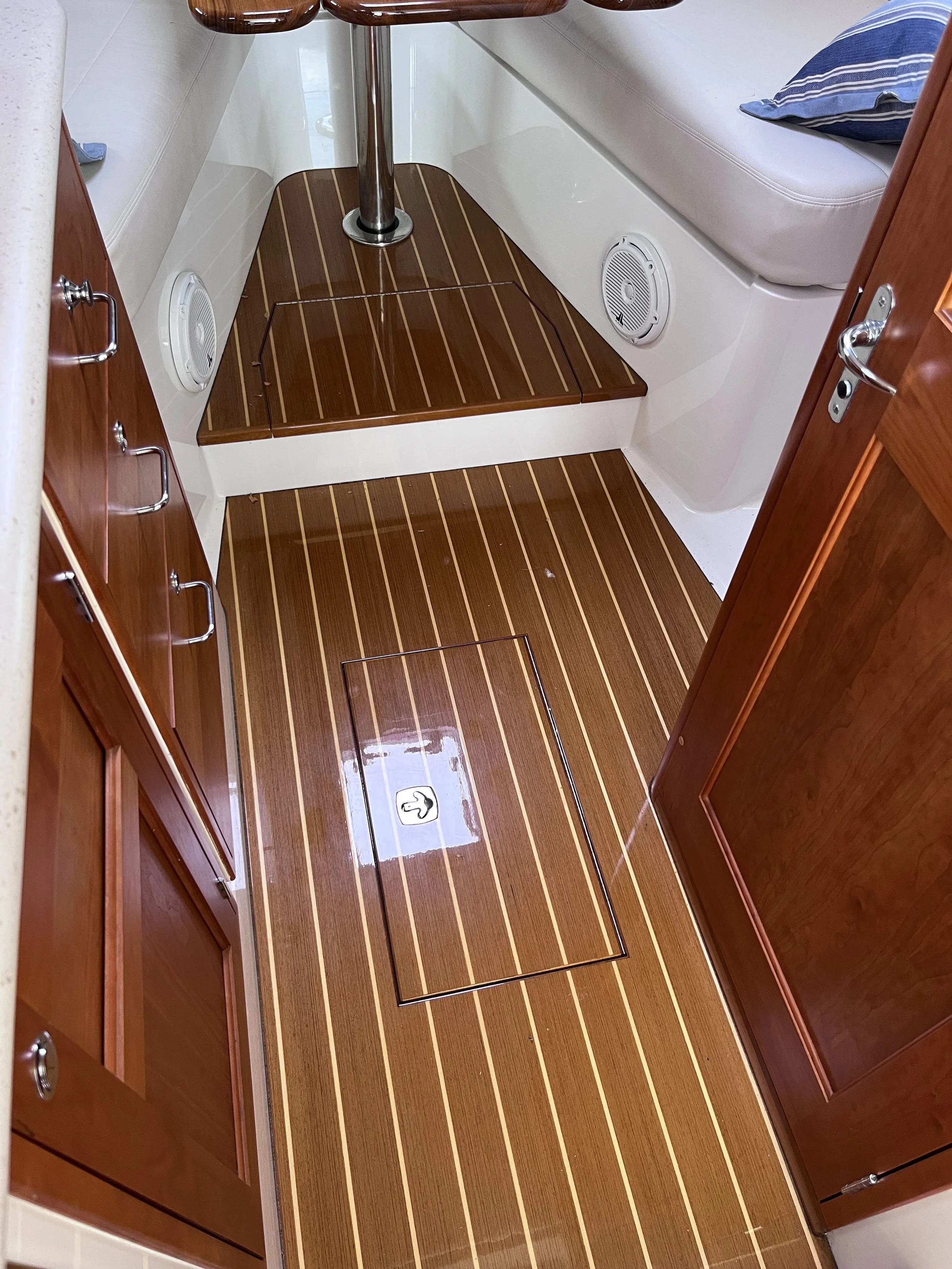 The image shows the interior of a boat's bathroom or changing area with wood textured flooring, built-in storage cabinets on the left, a bench seat with a cushion on the right, and two circular speakers on the wall. A hatch door with a handle is visi