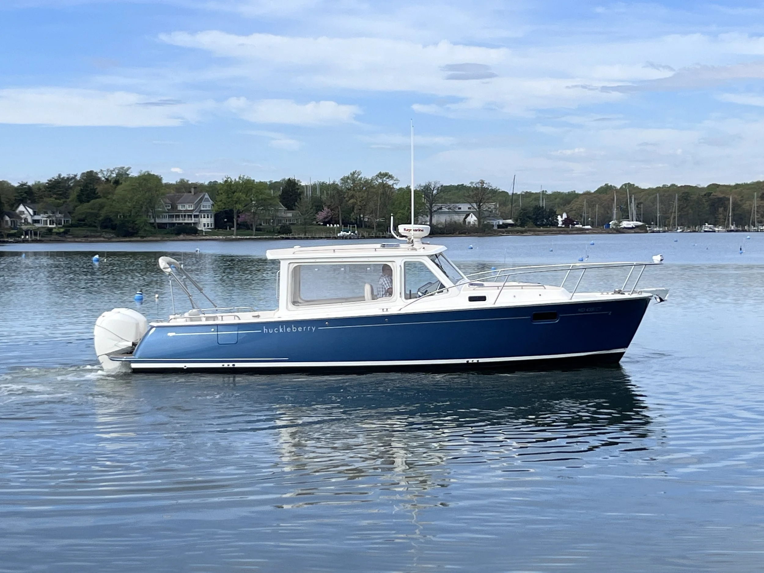 A blue and white boat with the name 'huckleberry' on the side, floating on calm water near a harbor with houses and sailboats in the background, under a partly cloudy sky.