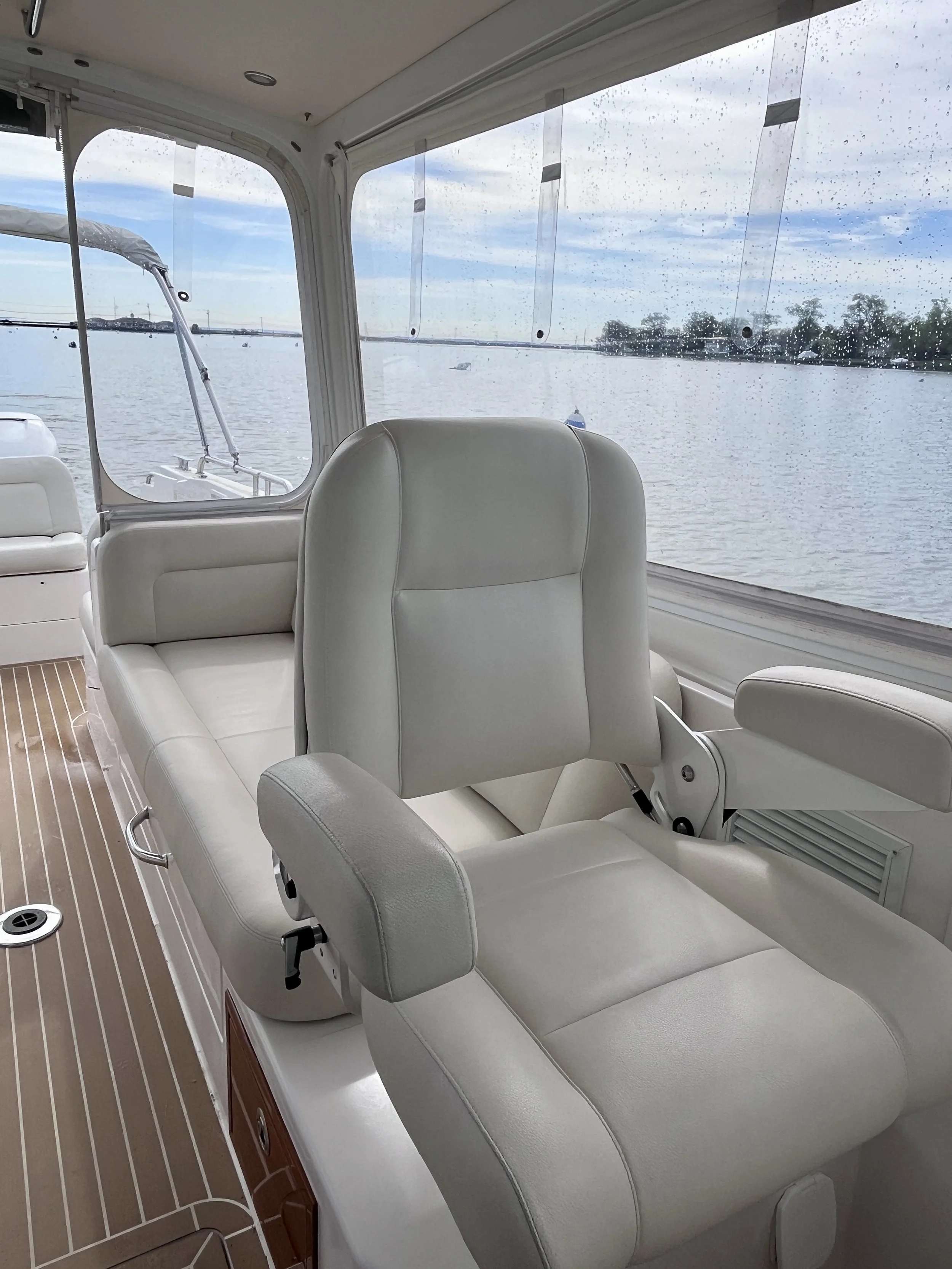 Interior of a boat with white leather seats and a large window showing water and boats outside, rain droplets on the window.