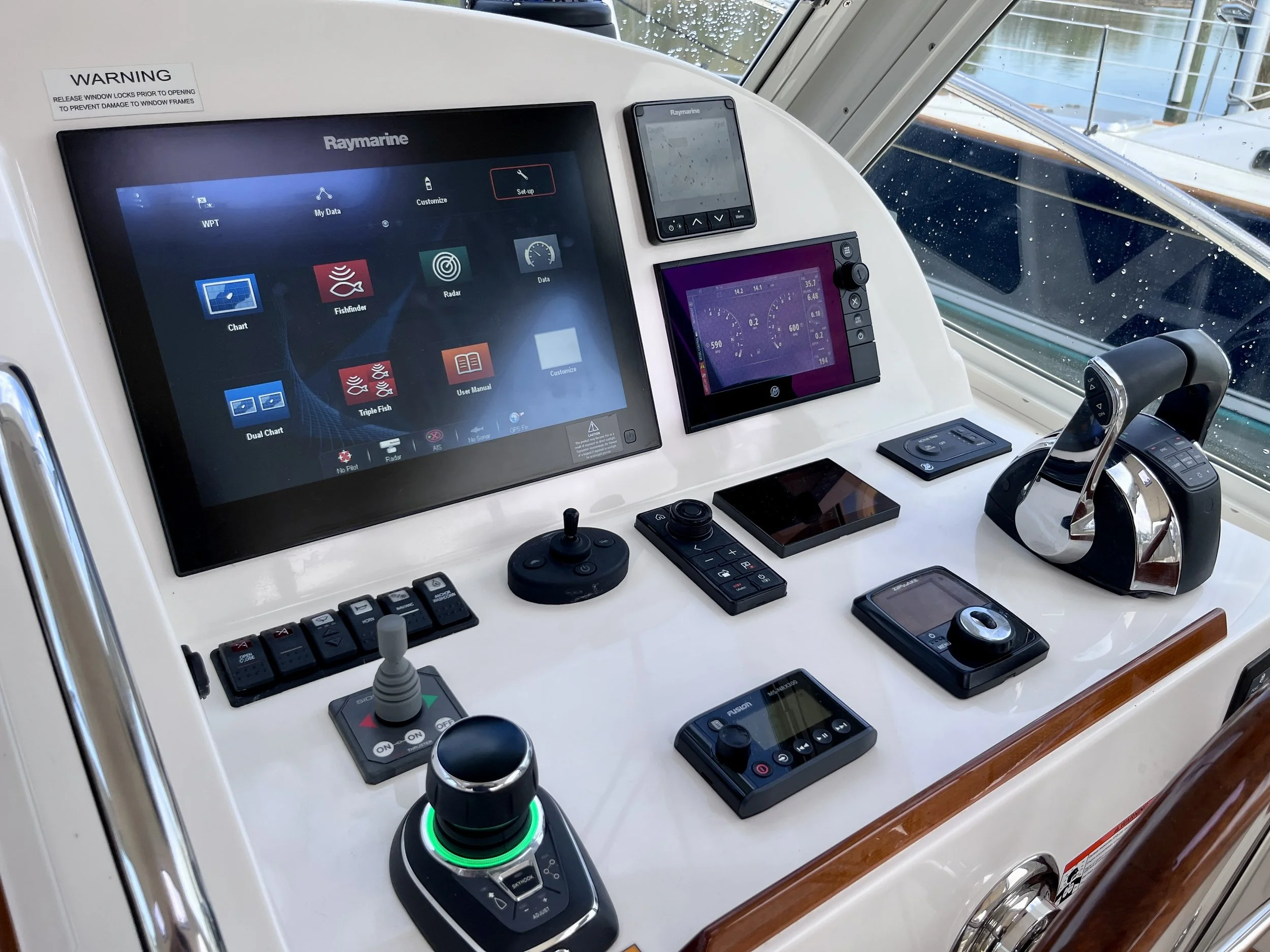 A boat cockpit with multiple electronic navigation and control screens, including a large Raymarine display, a smaller Raymarine device, and various control buttons, switches, and a joystick, with a partly rain-spattered window showing water and dock