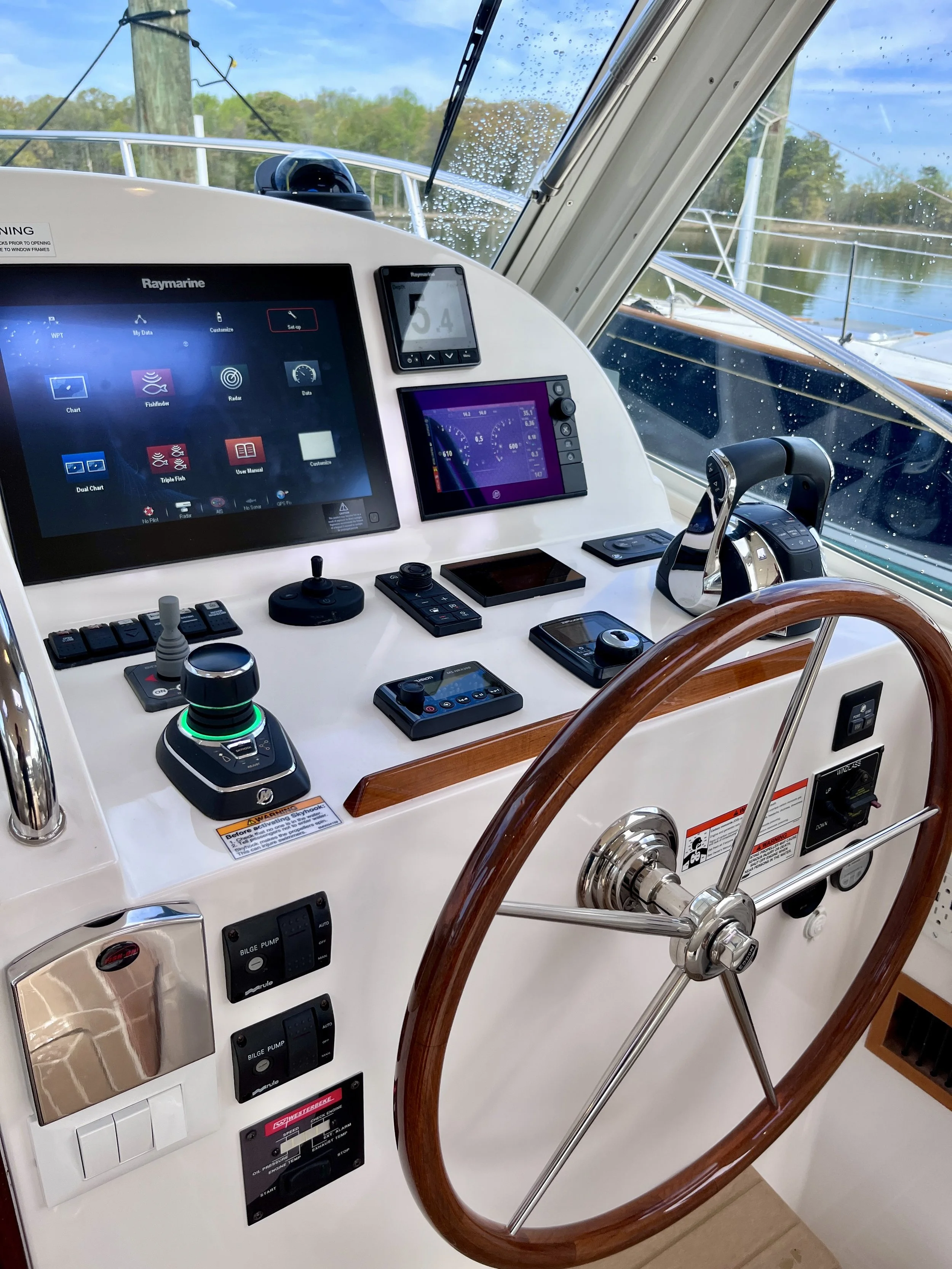 The helm station of a boat with multiple electronic navigation and control screens, a wooden steering wheel, and various switches and dials.