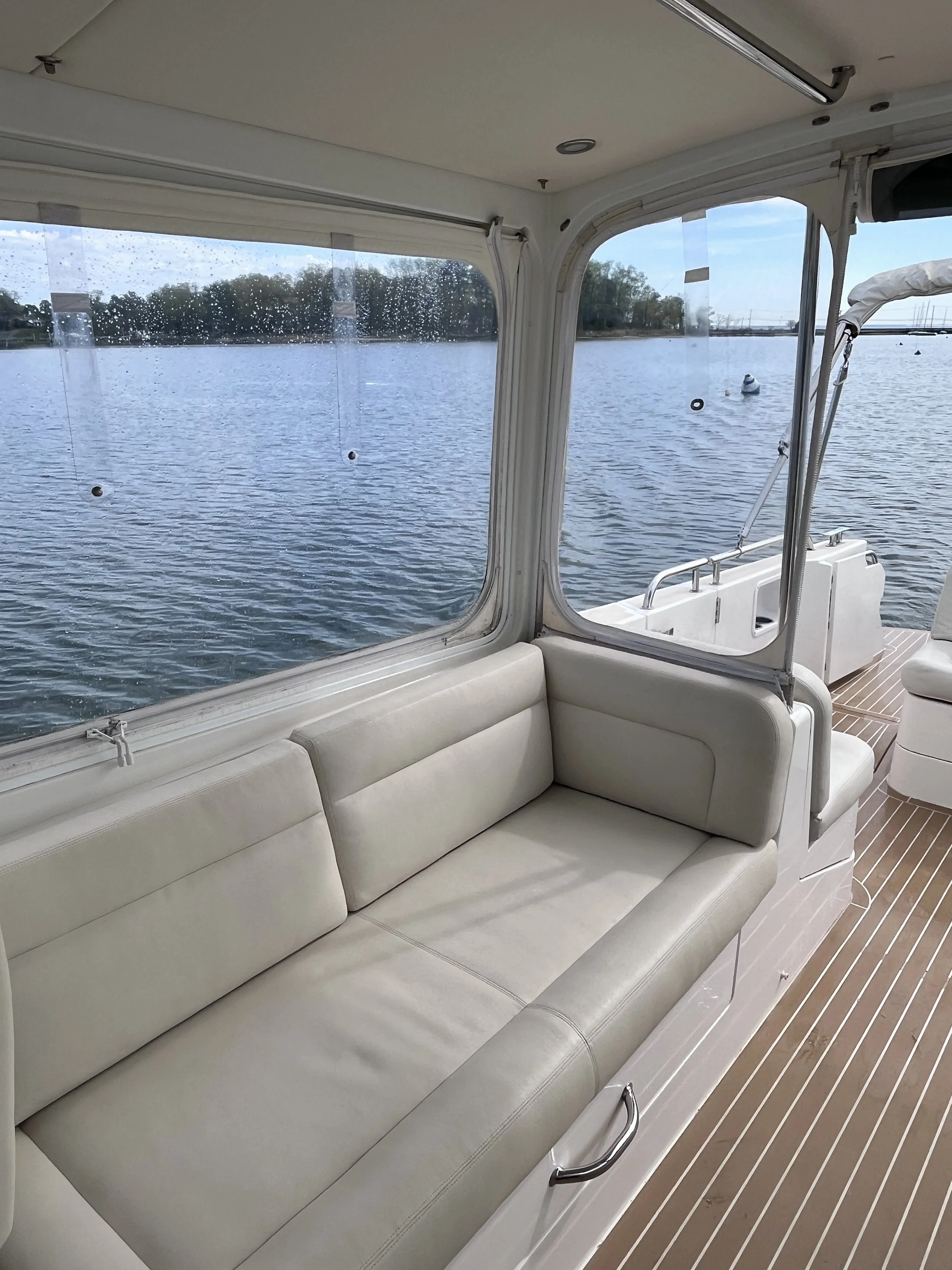 Interior view of a boat with white cushioned seating along the side, large windows showing water and trees outside, and a wooden deck floor.