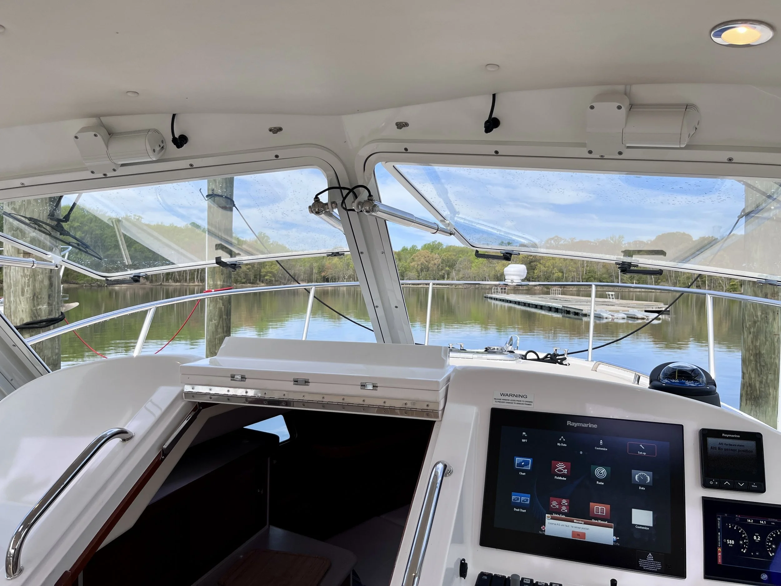 View from inside a boat's helm station, showing navigation and control screens, with a view of the water and trees outside through the windshield.