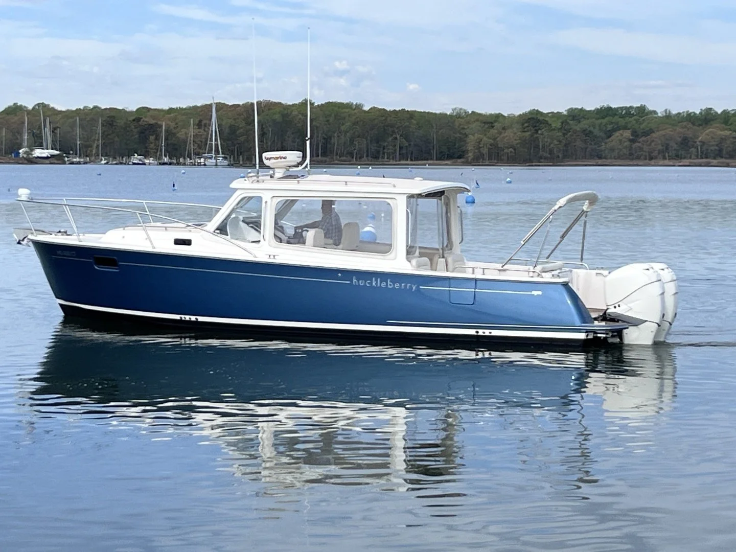 A blue and white motorboat named 'huckleberry' on calm water with a shoreline and trees in the background.