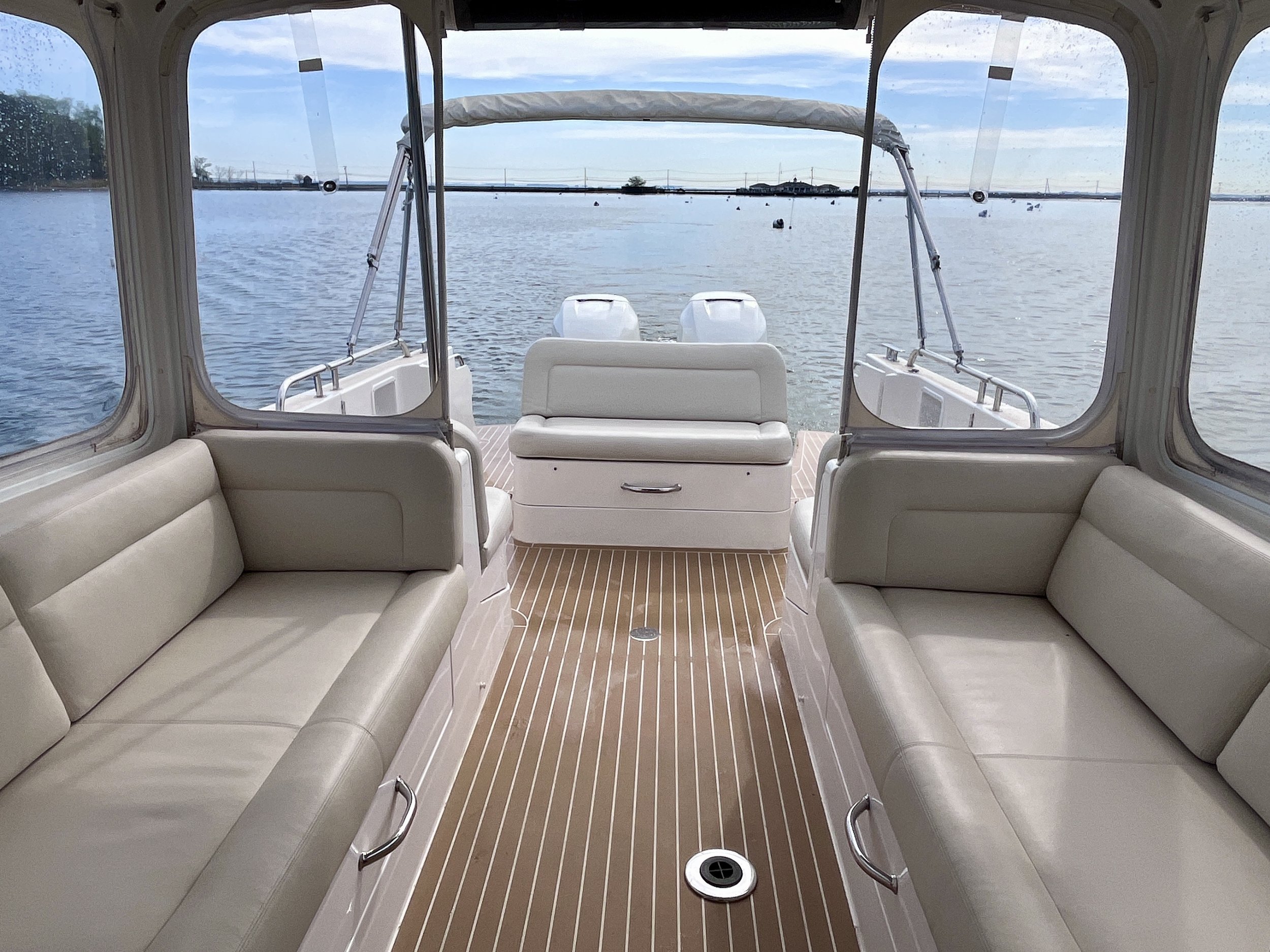 View from inside a boat showing cream-colored cushioned seating along the sides, a cushioned bench at the back, and a view of water with small boats and distant shoreline through clear windows.