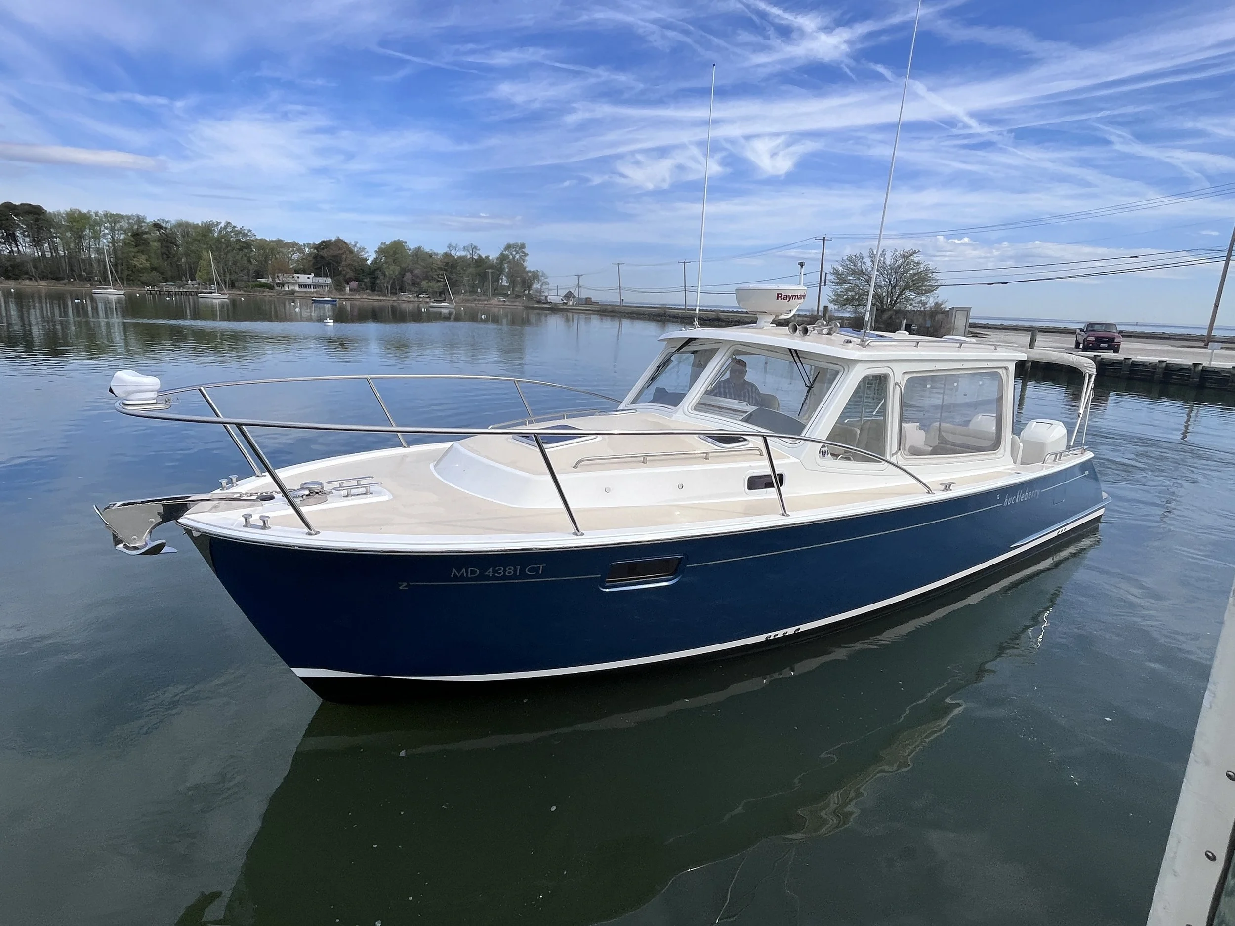 A small white and blue motorboat floating on calm water with reflections. In the background, there is a shoreline with trees, houses, boats, and a cloudy blue sky.