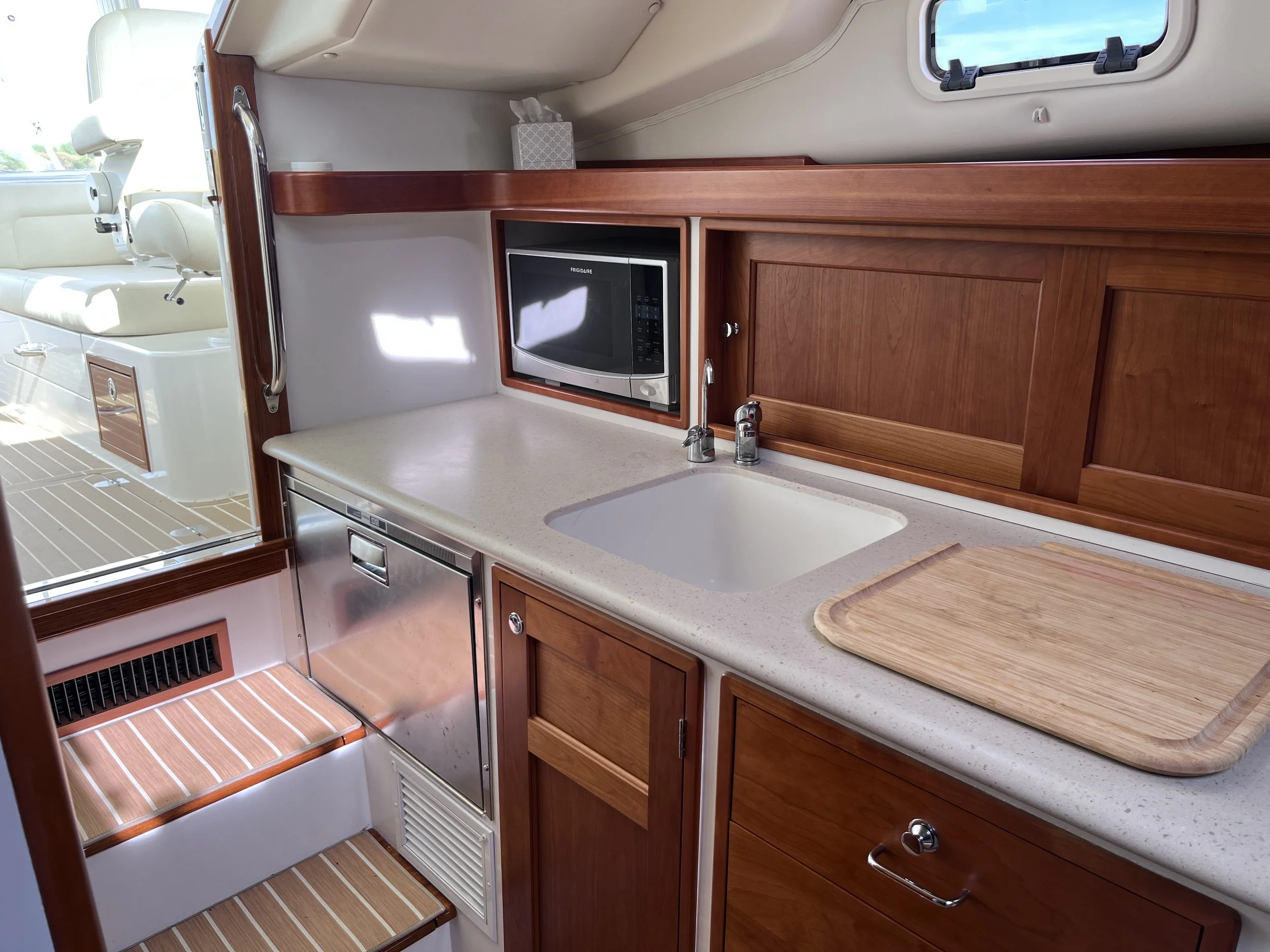 Interior of a boat kitchen with a countertop, a microwave oven, wooden cabinets, a sink, and a window showing the sky outside.