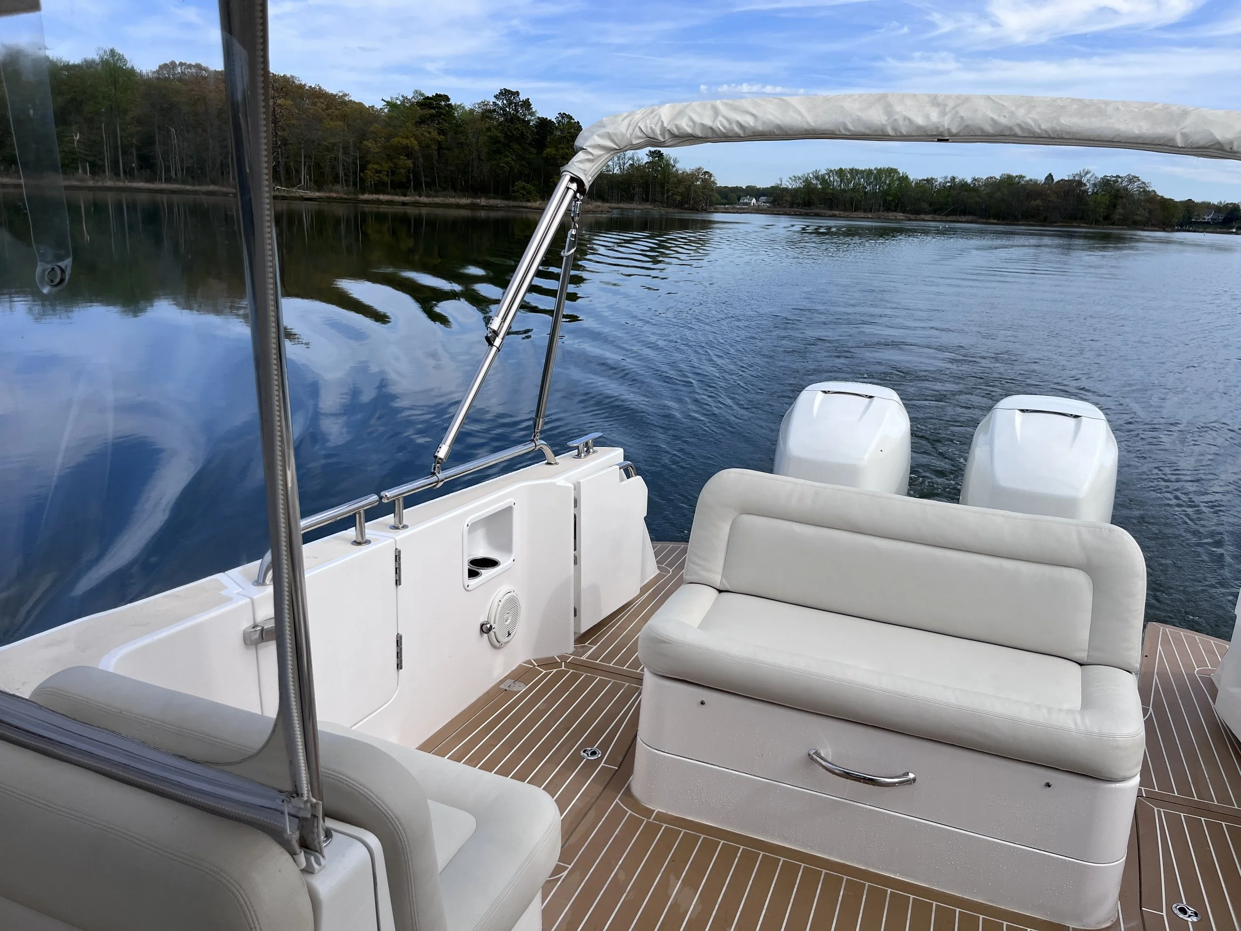View of a boat's deck with white seating and outboard motors, overlooking a calm river with trees and houses in the distance under a partly cloudy sky.