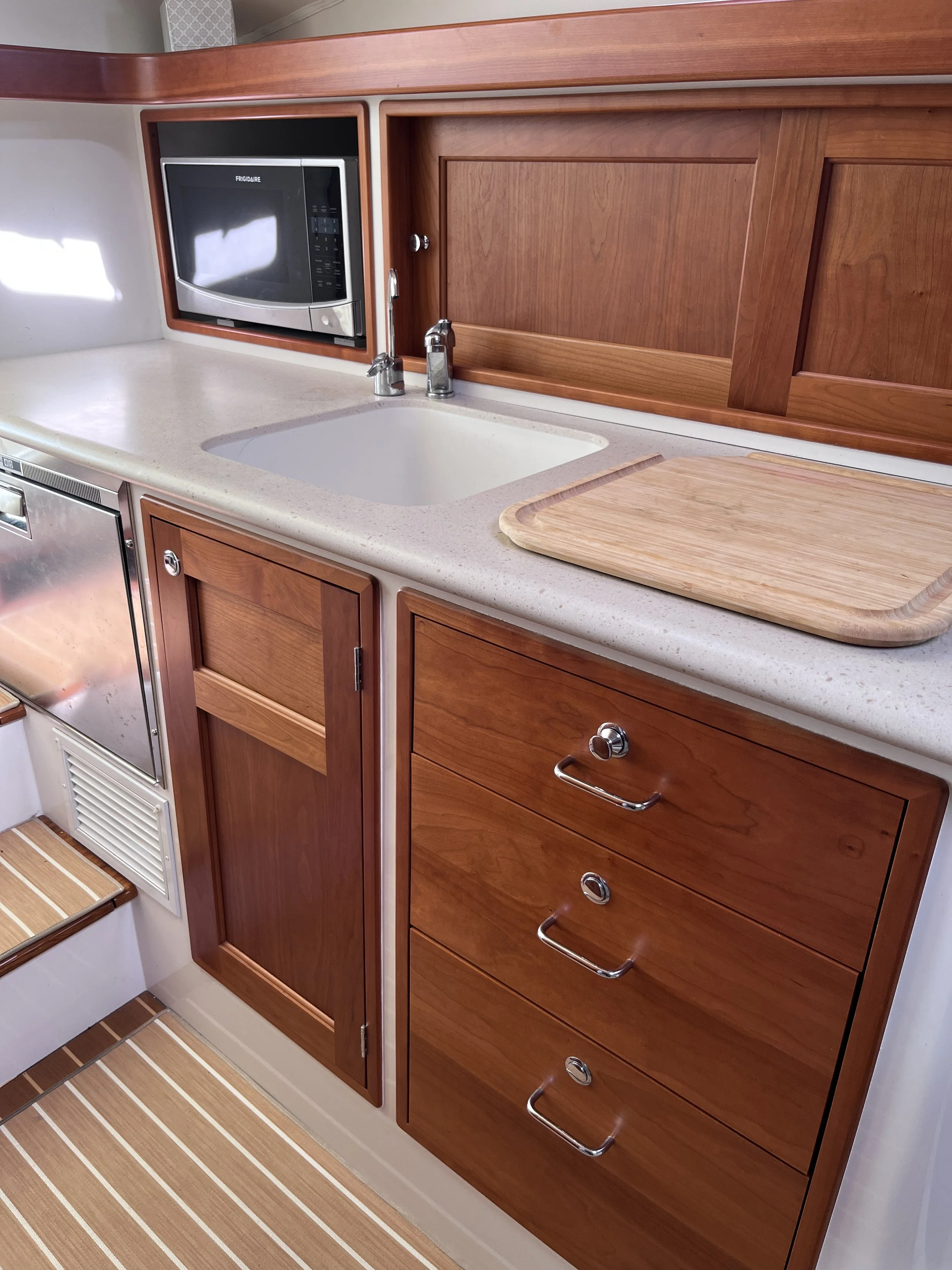 A kitchen with wooden cabinets, a microwave, a sink with a chrome faucet, and a wooden cutting board on the countertop.