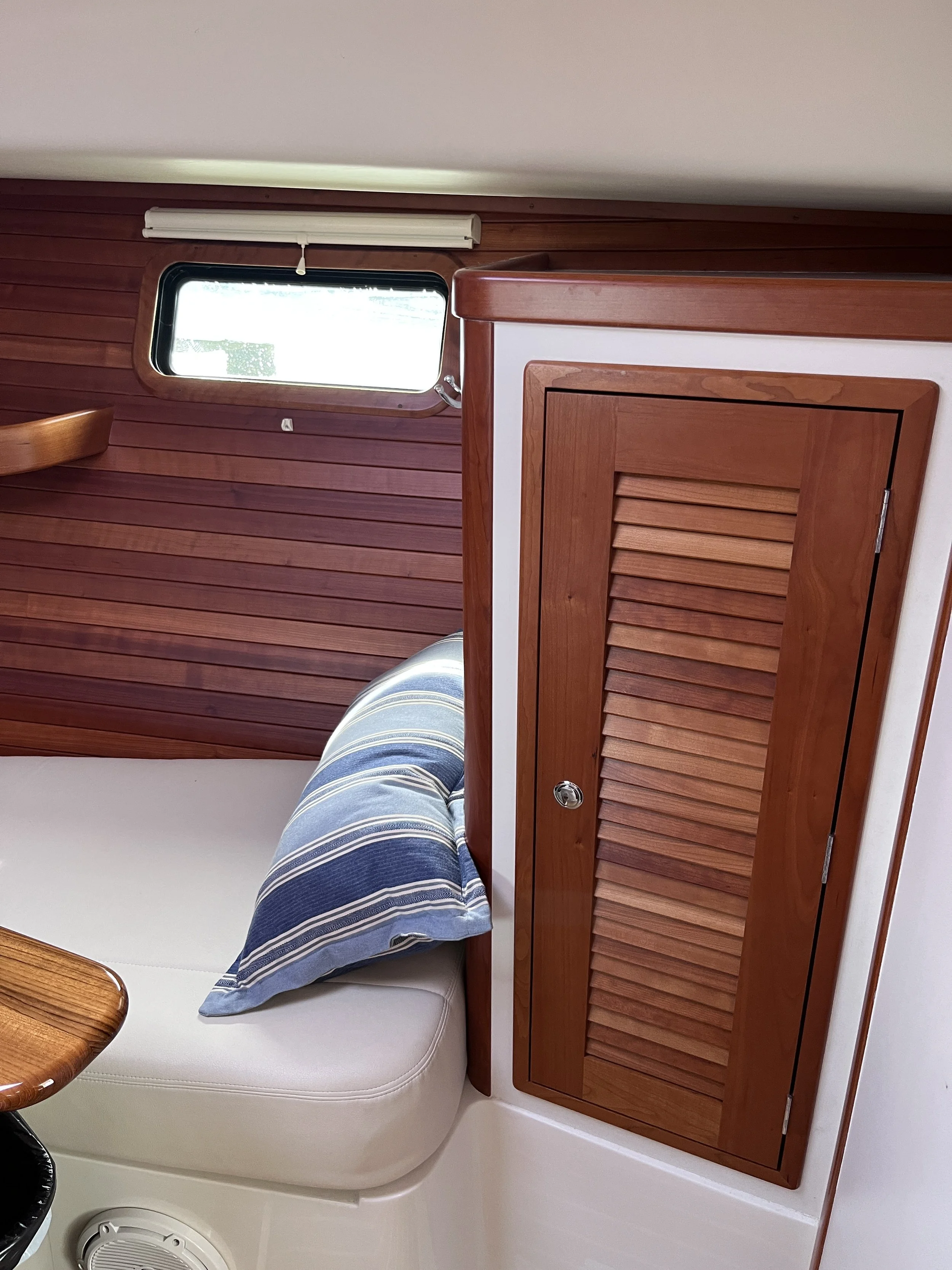 Interior of a boat with a small window, wooden cabinetry, a white cushioned seat with a blue and white striped pillow, and part of a table visible.