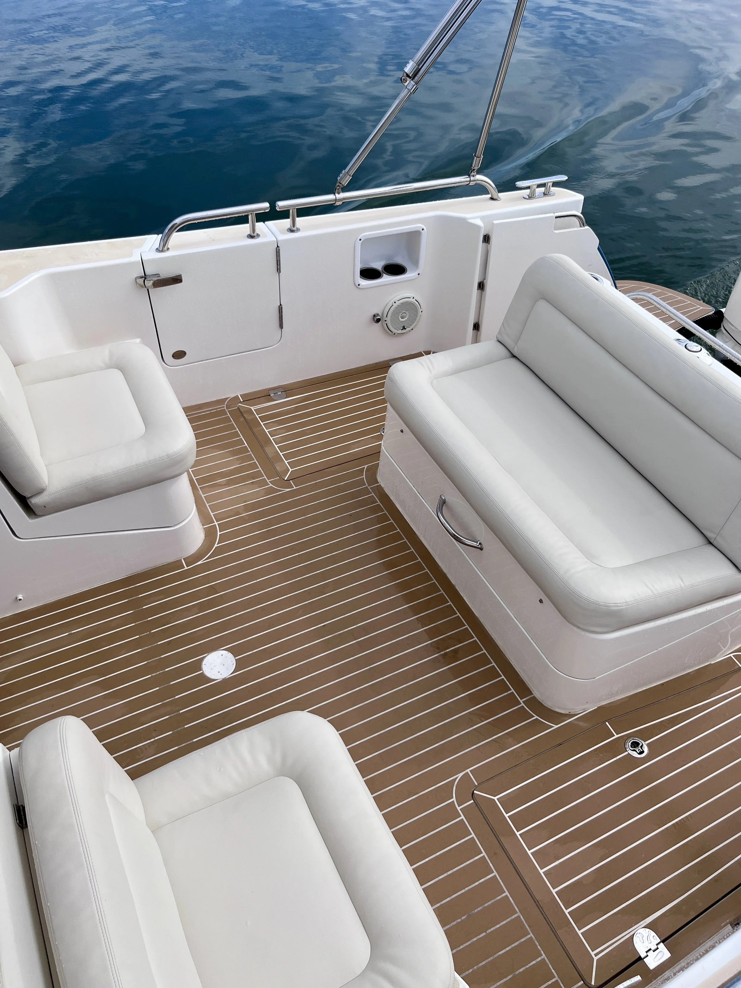 View of the deck on a boat, featuring white cushioned seats, stainless steel rails, and a wooden floor with white lines. Water is visible in the background.