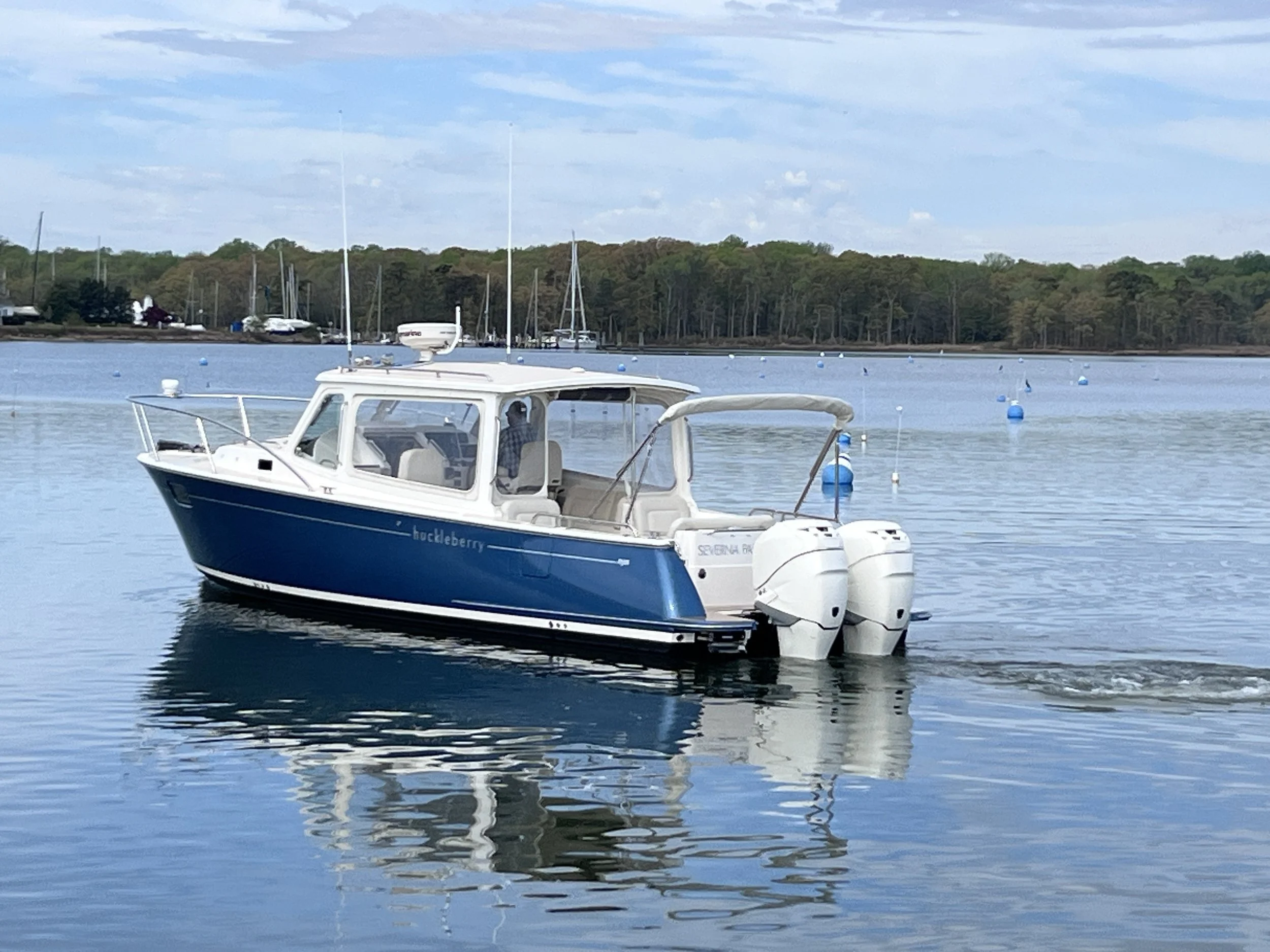 A small blue and white motorboat named 'Buckeleberry' floating on calm water with a man sitting inside, surrounded by boats and a shoreline with trees in the background.