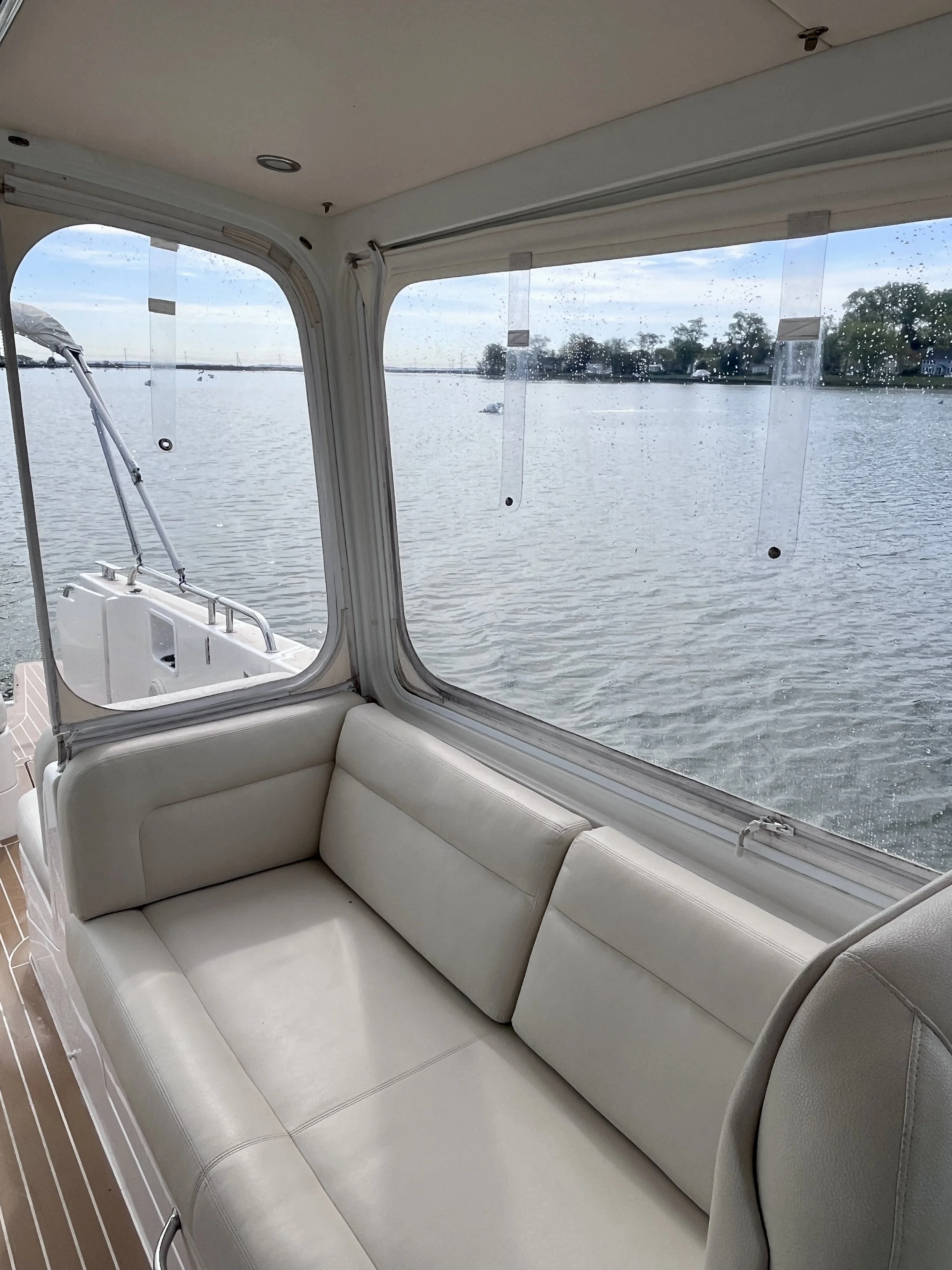 Interior view of a boat with a white cushioned seating area and windows showing water and trees outside.