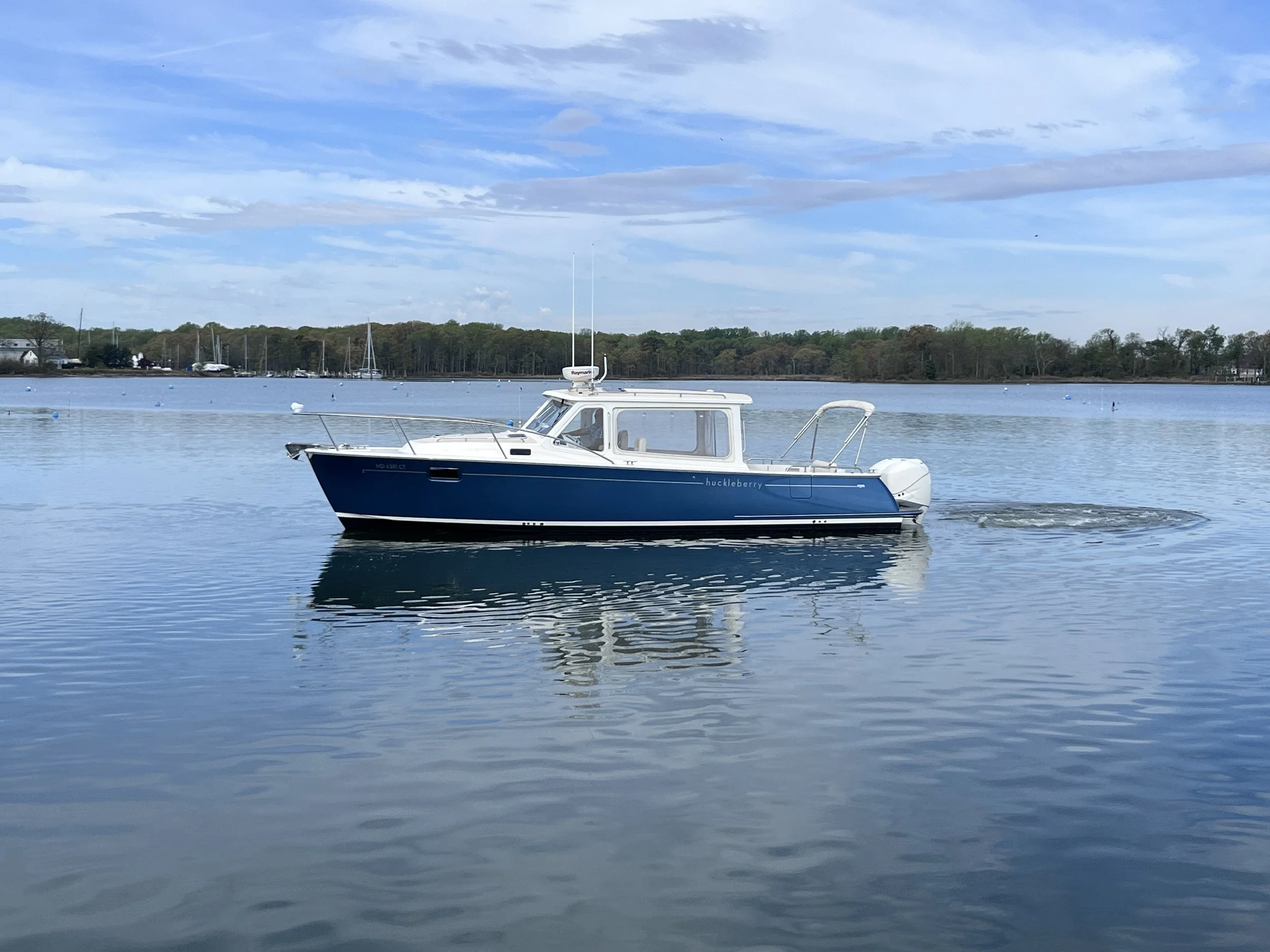 A boat named 'huckleberry' on a body of water with trees and boats in the background under a partly cloudy sky.