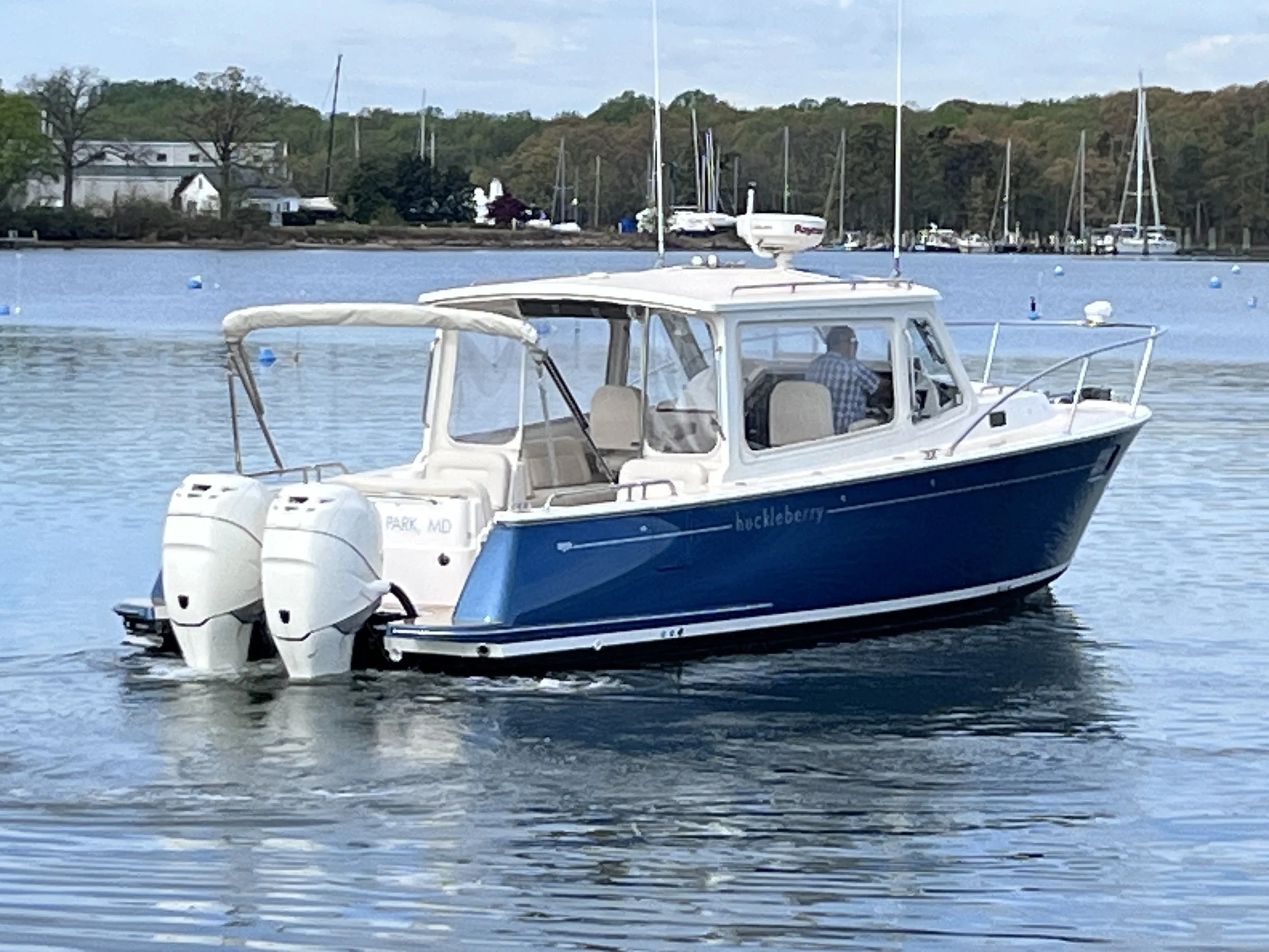 A motorboat with twin outboard motors moving across a body of water near a marina, with sailboats docked, trees, and houses in the background.