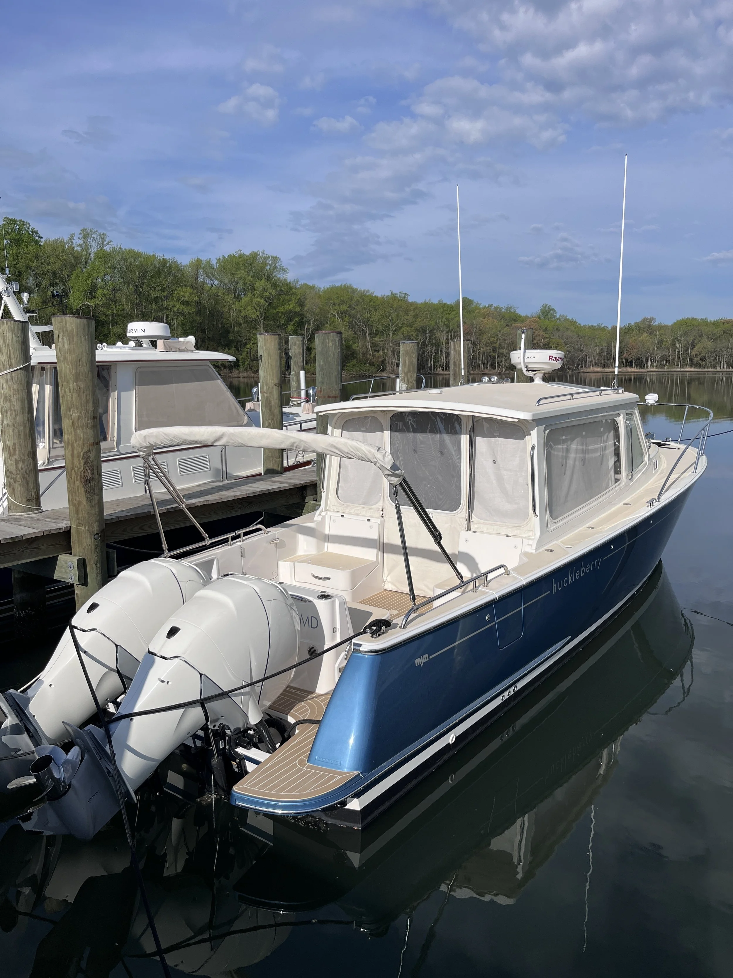 A boat docked at a marina with tall wooden posts and a dock. The boat has twin outboard motors, a white cabin, and is named 'Huckleberry.' The background shows a calm body of water and a tree-lined shoreline under a partly cloudy sky.