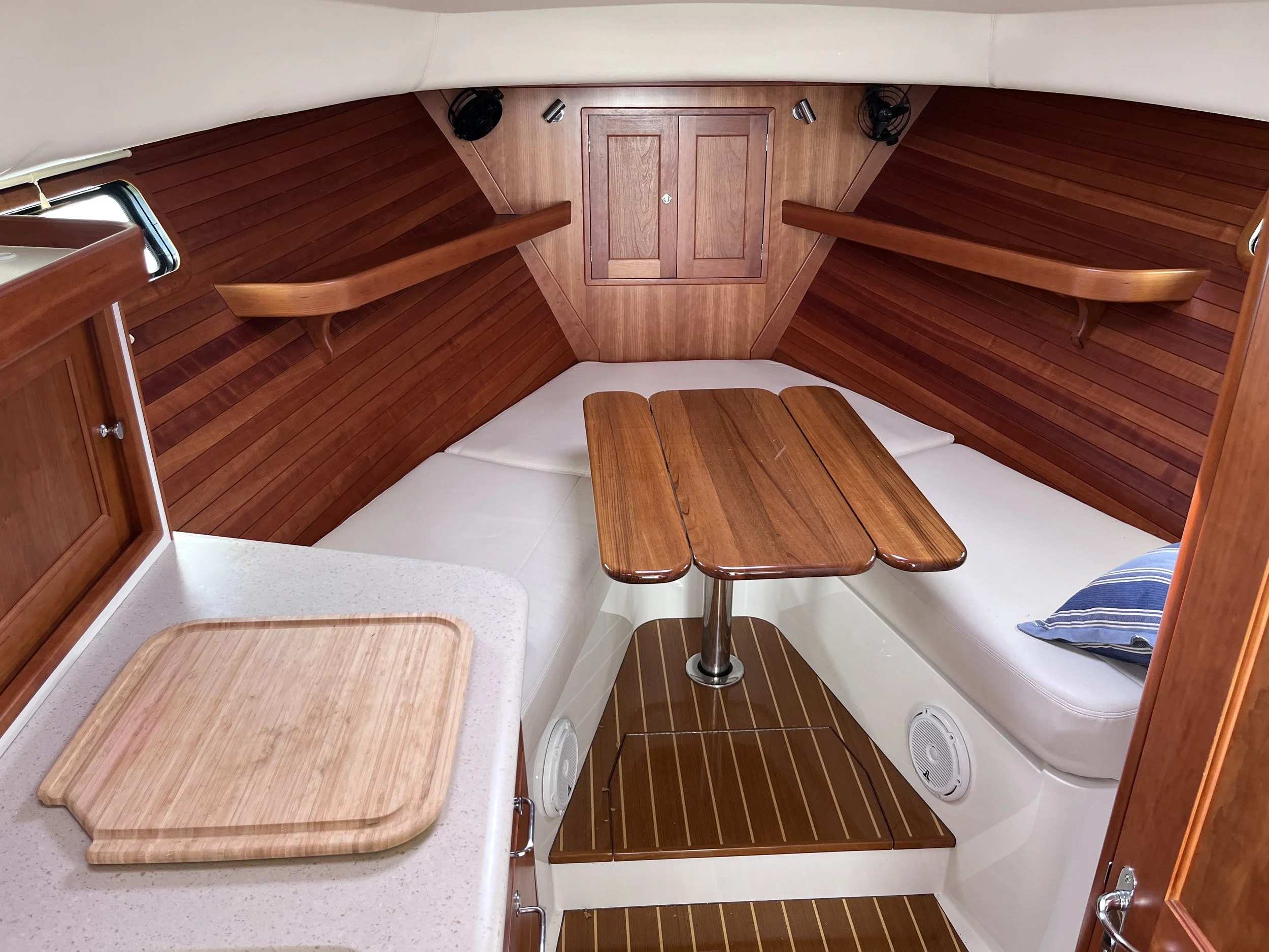 Interior view of a boat cabin with a V-shaped wooden seating area, a small wooden table in the center, wooden panels on the walls, and various ventilation fans.