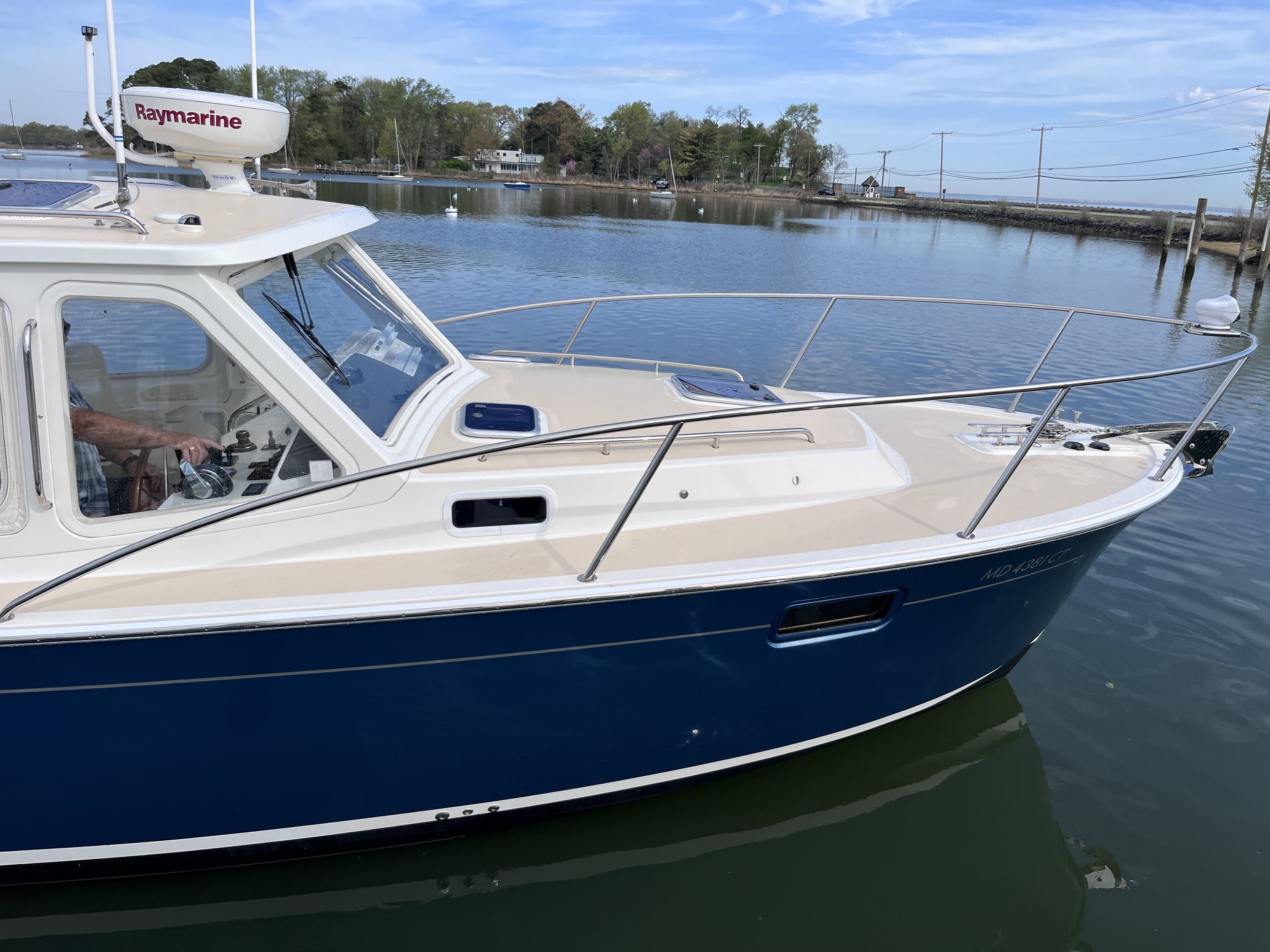 A white motorboat docked at a marina with calm water. In the background, there are trees, a building, and power lines under a partly cloudy sky.