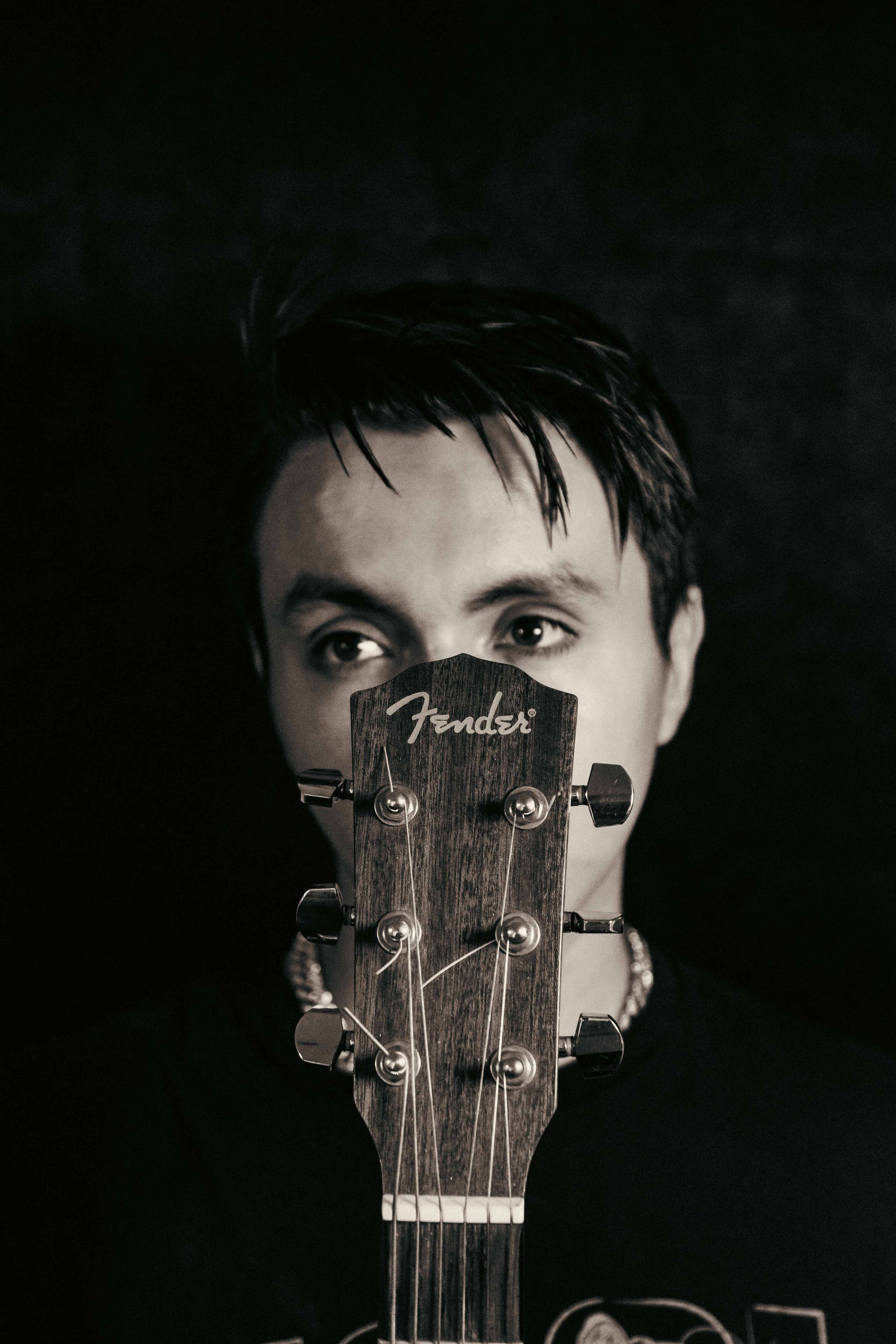 A close-up black and white photo of a person's face, with the head partially obscured by a guitar's headstock covering the nose and mouth. The person's eyes are visible, looking upward, with wet or styled dark hair. The guitar brand Fender is visible on the headstock.