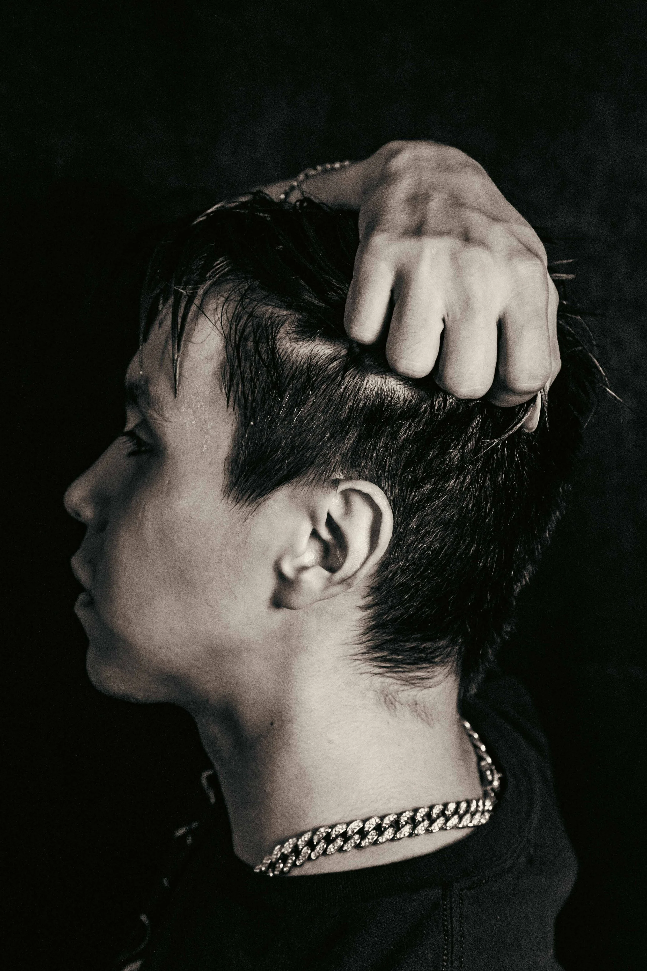 Black and white photo of a person with short hair, wearing a chain necklace and a black top, with their hand on their head and their face turned to the side.