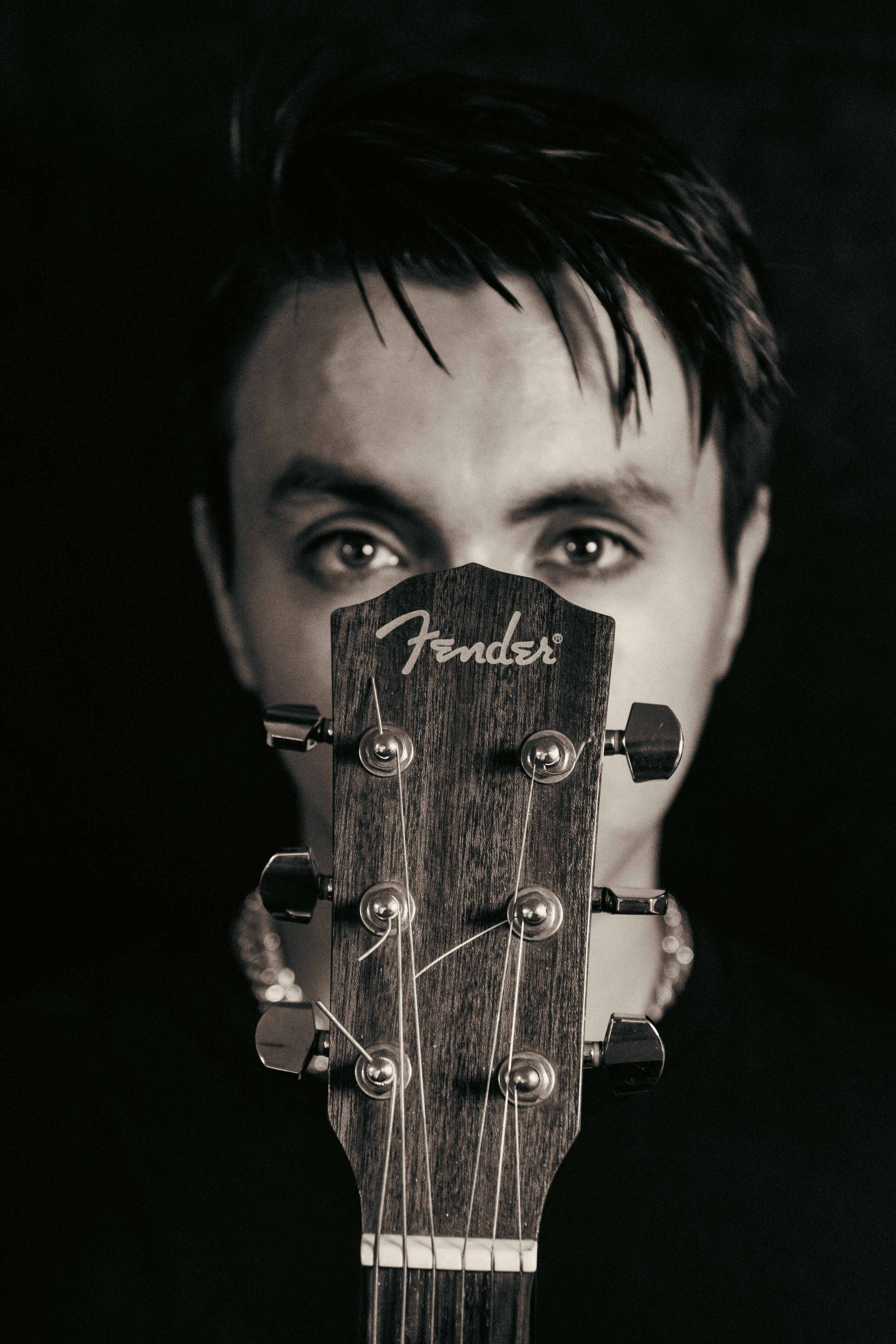 Close-up of a person's face with dark hair, looking through the headstock of a Fender guitar, which partially obscures their mouth and nose, in black and white.