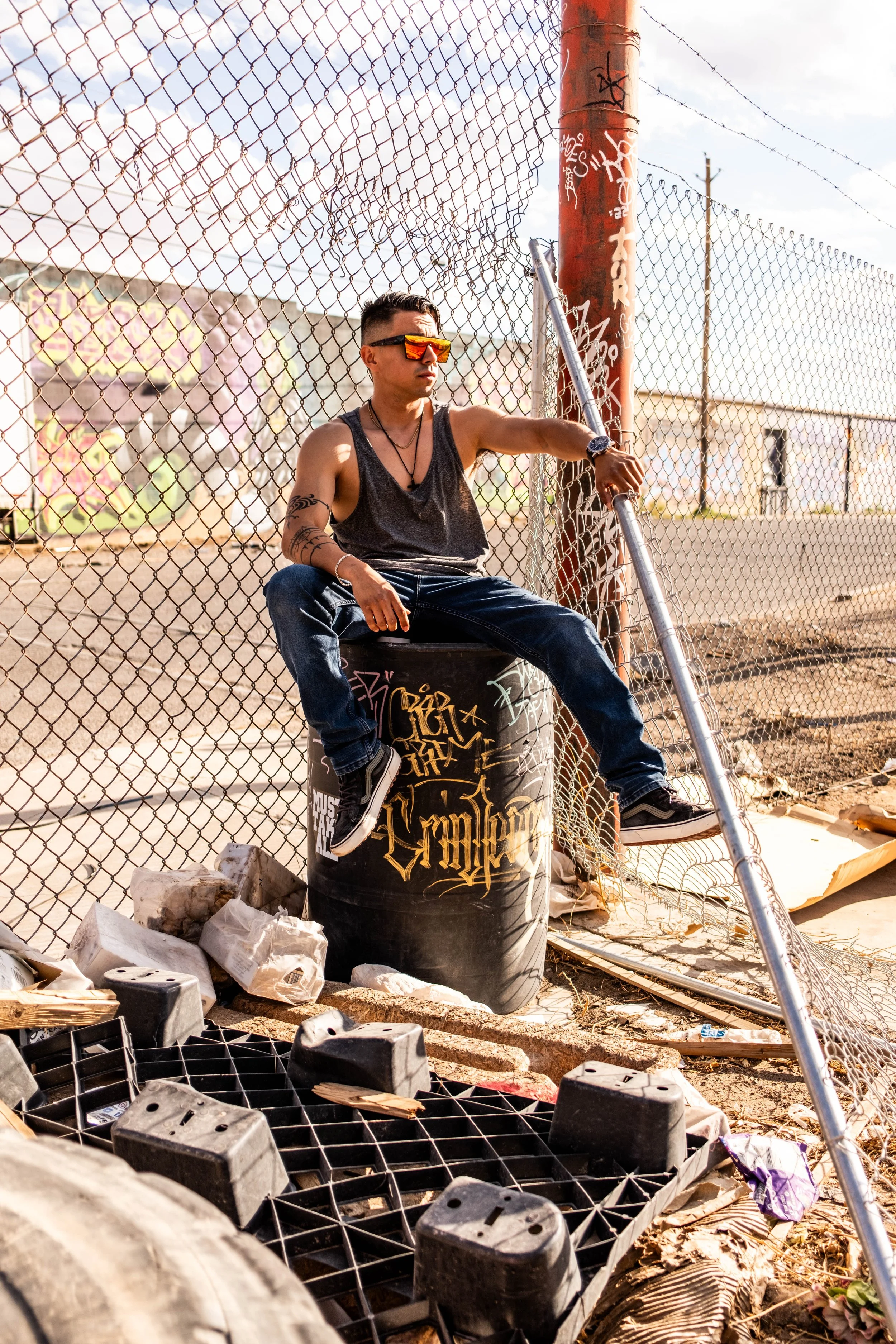 A young man with tattoos and sunglasses sitting on a trash can in front of a chain-link fence, with debris and rocks on the ground, in an urban setting.