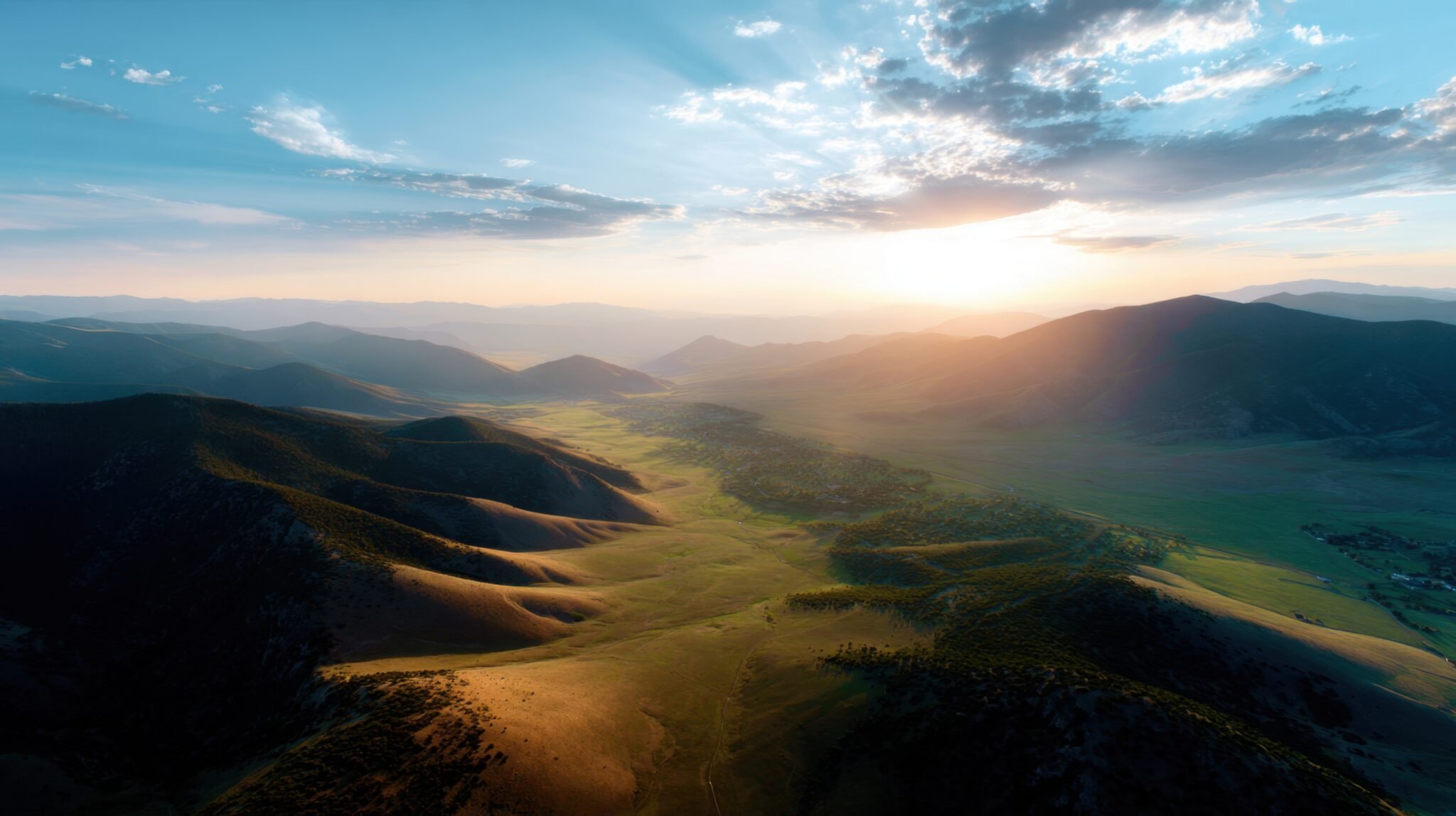A scenic landscape of rolling green hills and distant mountains under a partly cloudy sky with the sun setting or rising.