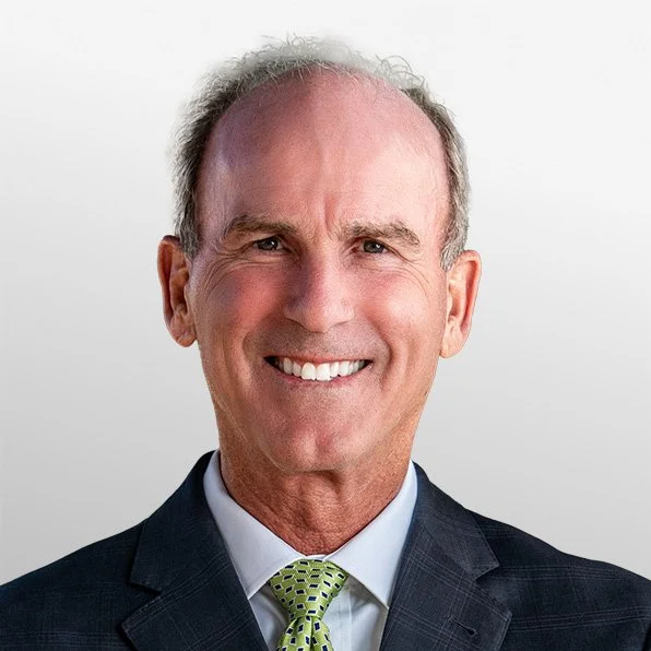 A middle-aged man with short gray hair, wearing a dark suit, white shirt, and a green patterned tie, smiling against a plain light background.