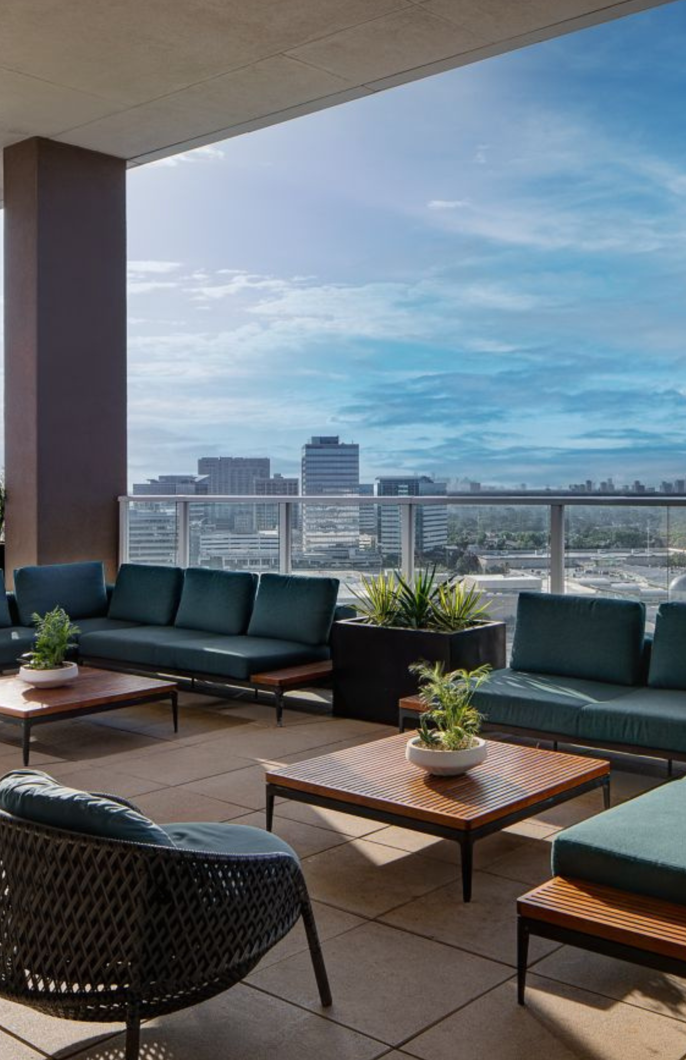 An outdoor rooftop lounge with modern teal sofas, wooden coffee tables with potted plants, overlooking a city skyline with tall buildings under a partly cloudy sky.