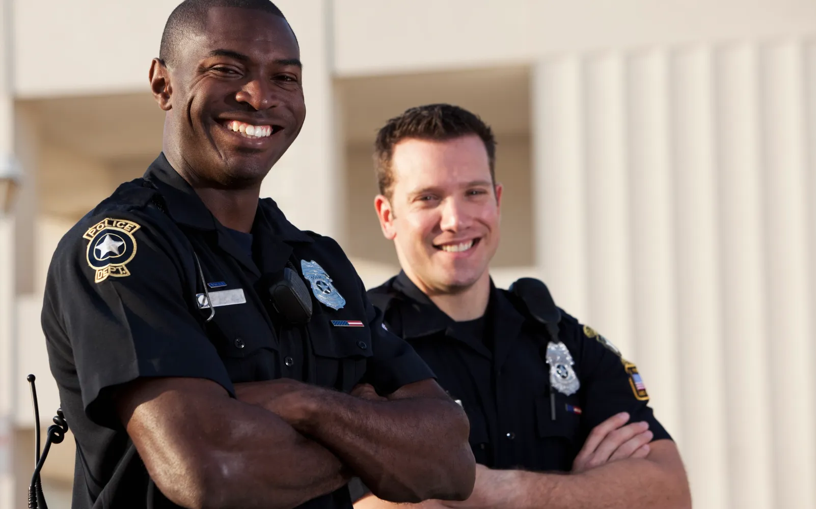 Two police officers standing outdoors, smiling with arms crossed, with white pillars in the background.