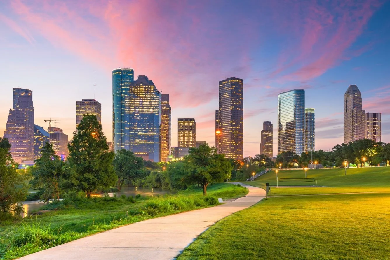 A city skyline at sunset with modern skyscrapers and a park in the foreground, including green grass, trees, and a paved walkway.