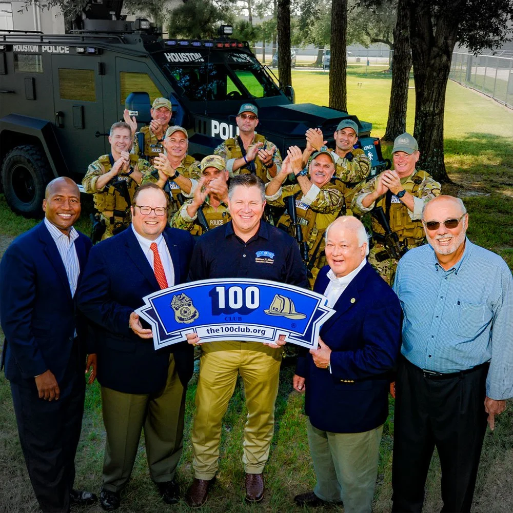 Group of people, including police officers and community members, holding a sign celebrating the 100th club, with a police vehicle in the background.