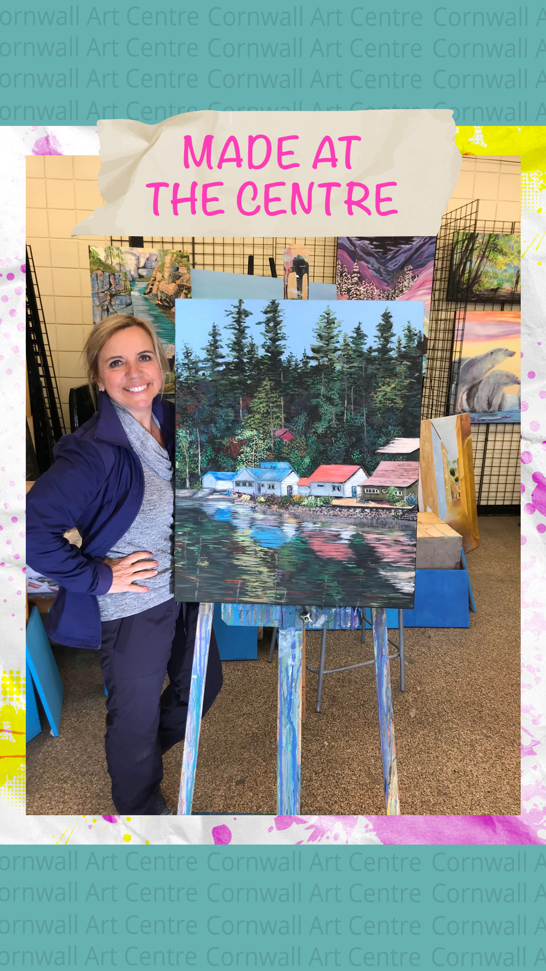 A woman standing next to an easel with a painting of a lakeside scene with small houses, tall trees, and reflections in the water. Artwork is displayed at an art center.