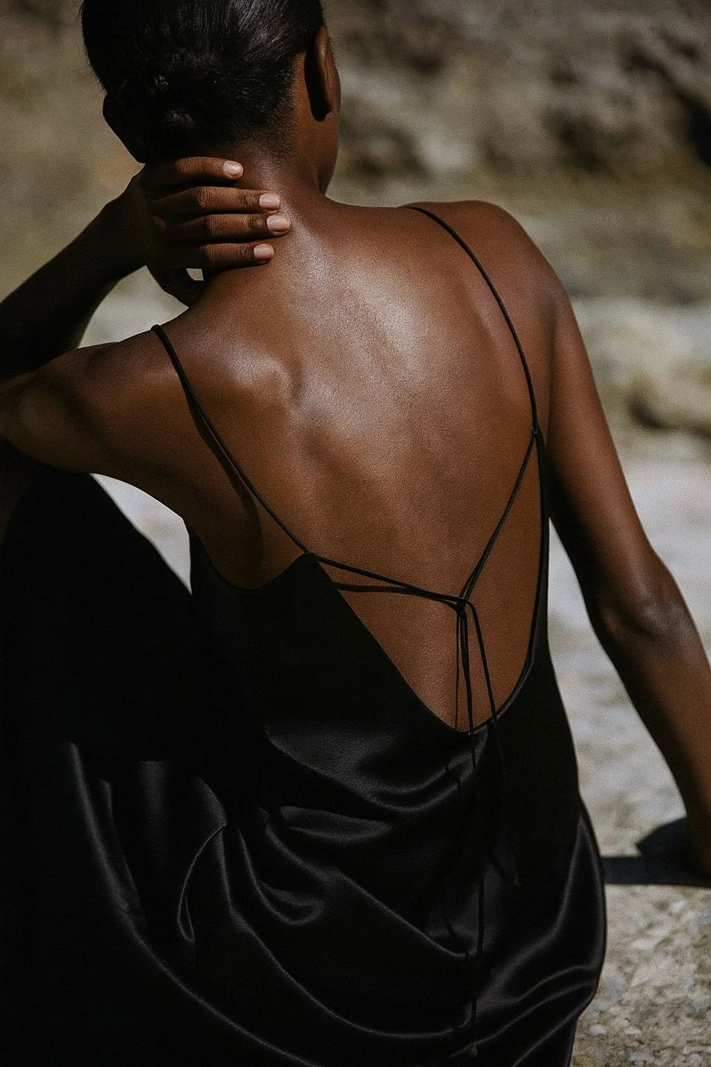 A woman with dark skin, wearing a black spaghetti strap dress, sitting outdoors with her back facing the camera, her left hand touching the back of her neck.