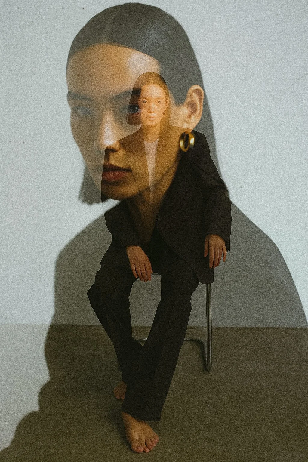 A creative photo where a woman in a black suit is seated on a chair, and her large, superimposed face creates a layered, artistic effect.