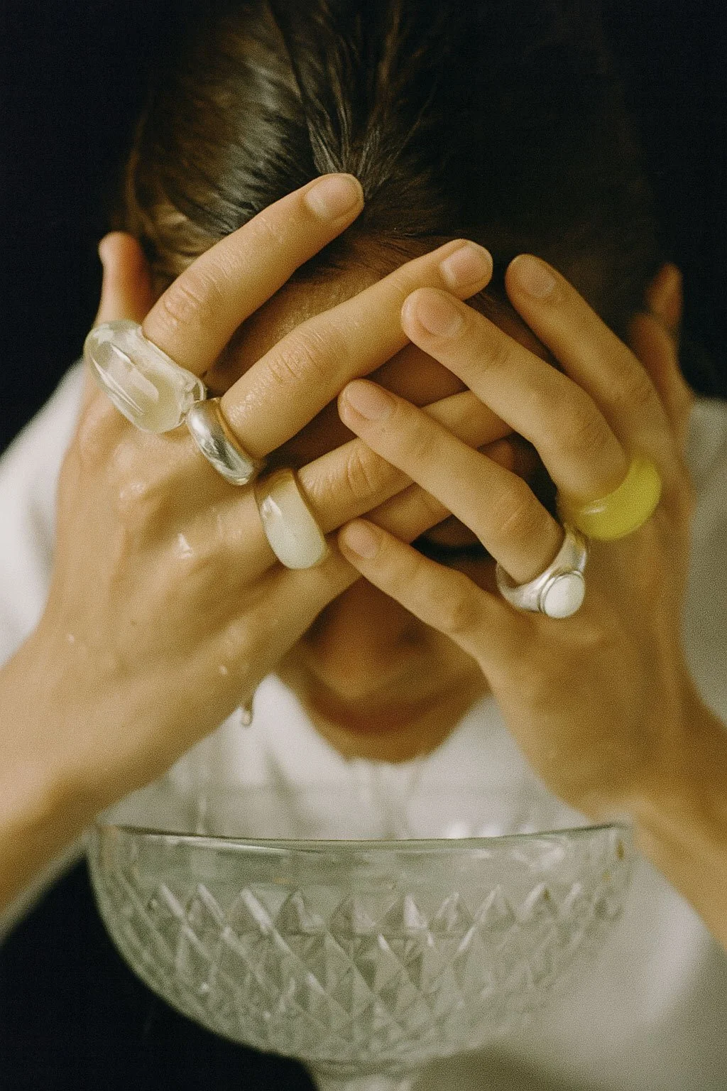 Person with dark hair covering face, hands with multiple rings, resting on forehead, in front of a patterned glass bowl.