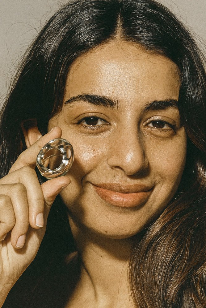 A woman with dark hair holding a glass object near her face, smiling gently, with a neutral background.