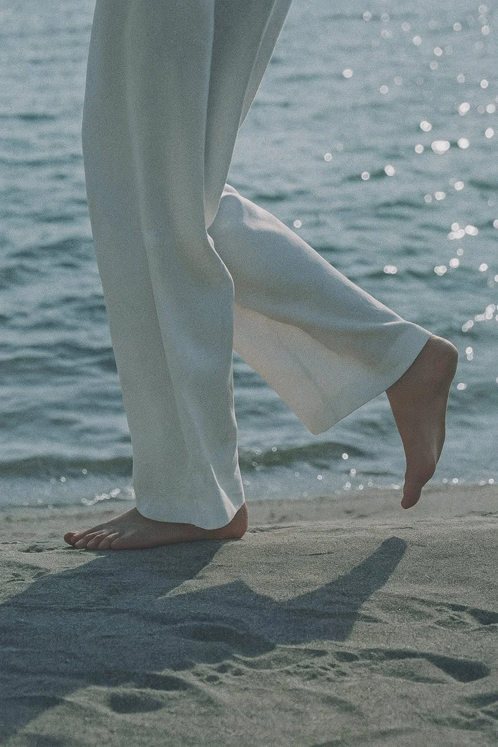 Person in white pants barefoot walking on a beach with water in the background