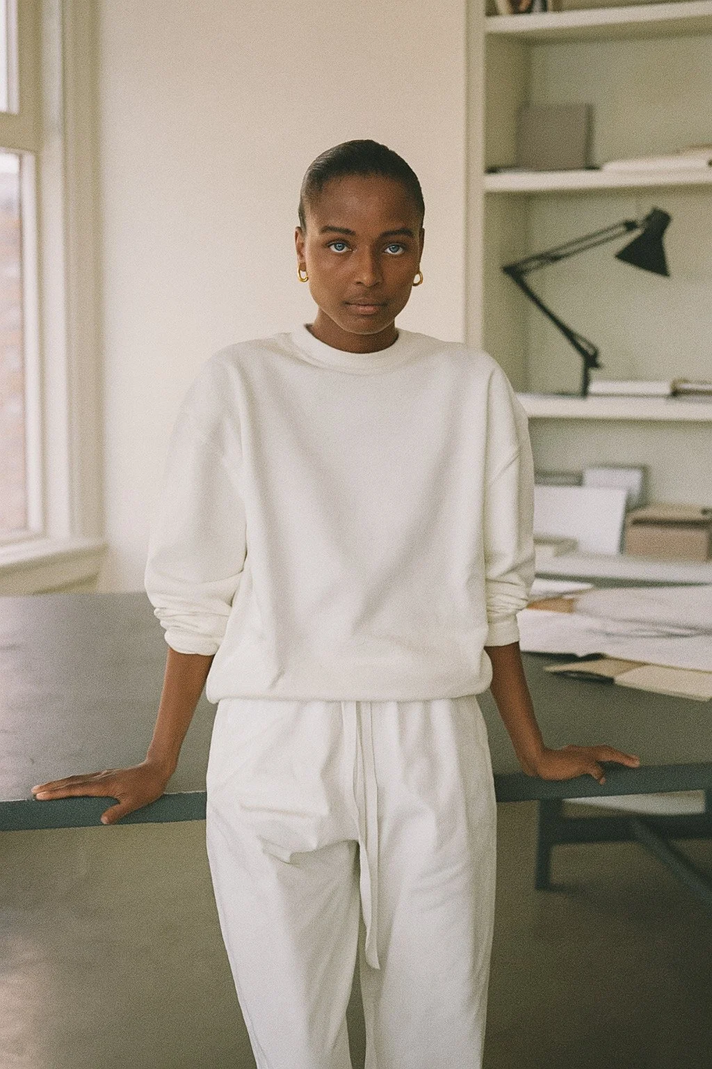 A woman with dark skin and braided hair, wearing a white sweatshirt and white pants, standing in a room with a desk and shelves behind her.