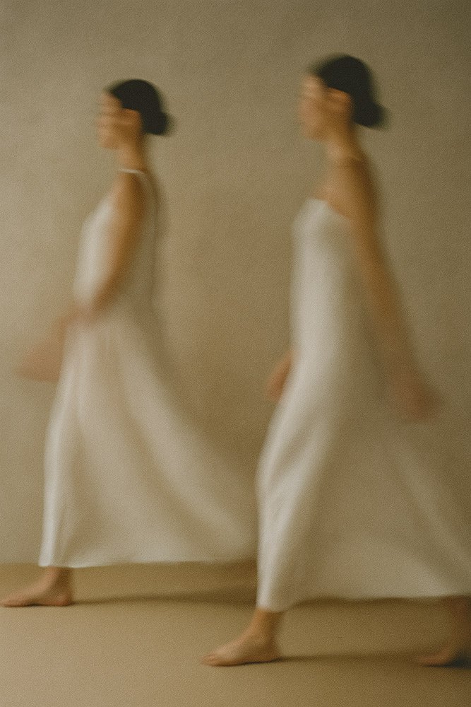Blurred photo of two women in white dresses walking barefoot against a plain background.