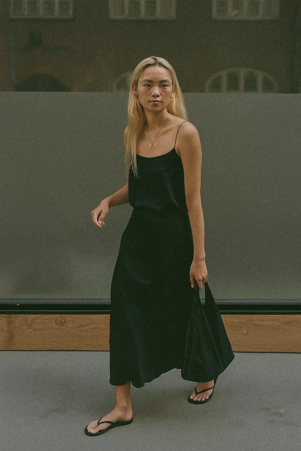 A woman with long blonde hair wearing a black spaghetti strap dress and flip-flops, walking in a dimly lit indoor space.