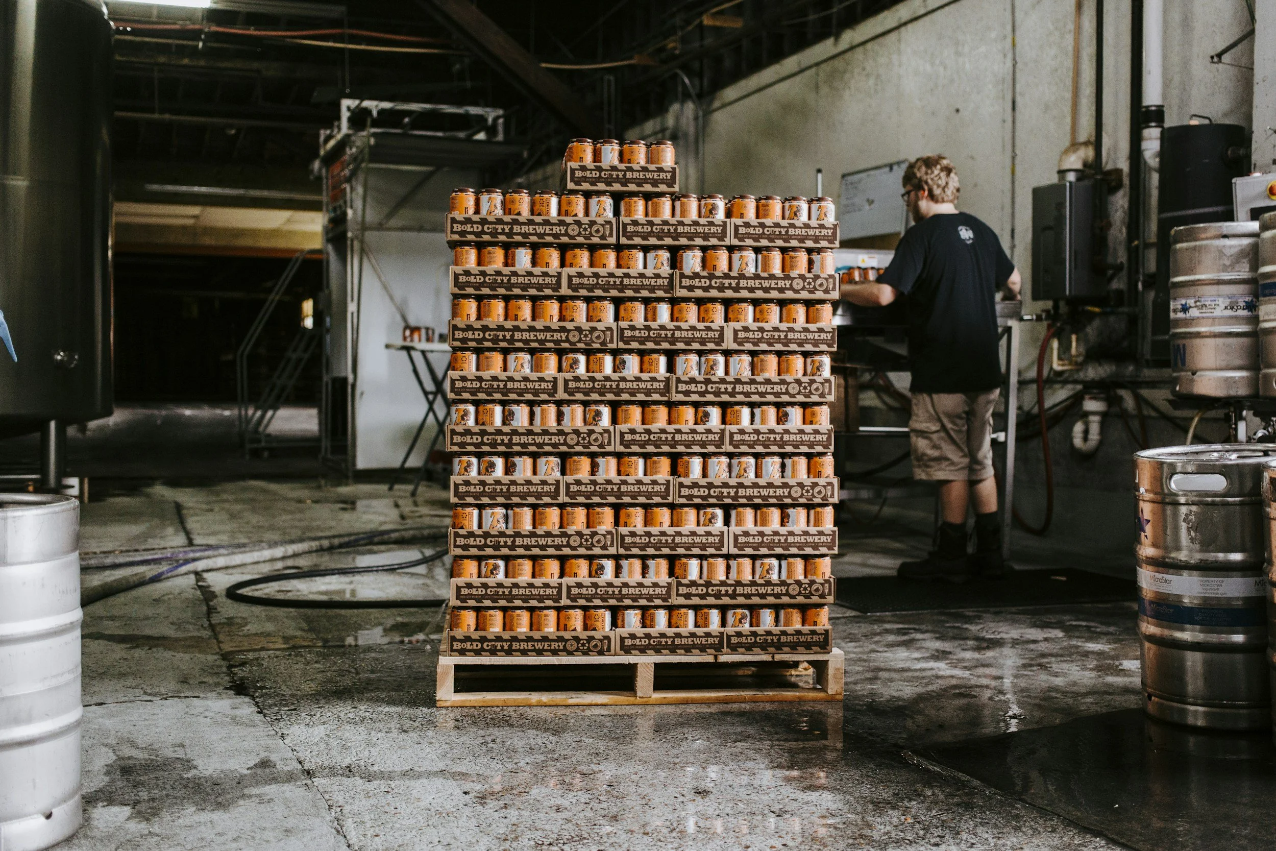 A tall pallet of cans labeled BOLD CITY BREWERY in an industrial setting with a person working in the background, surrounded by barrels and equipment.
