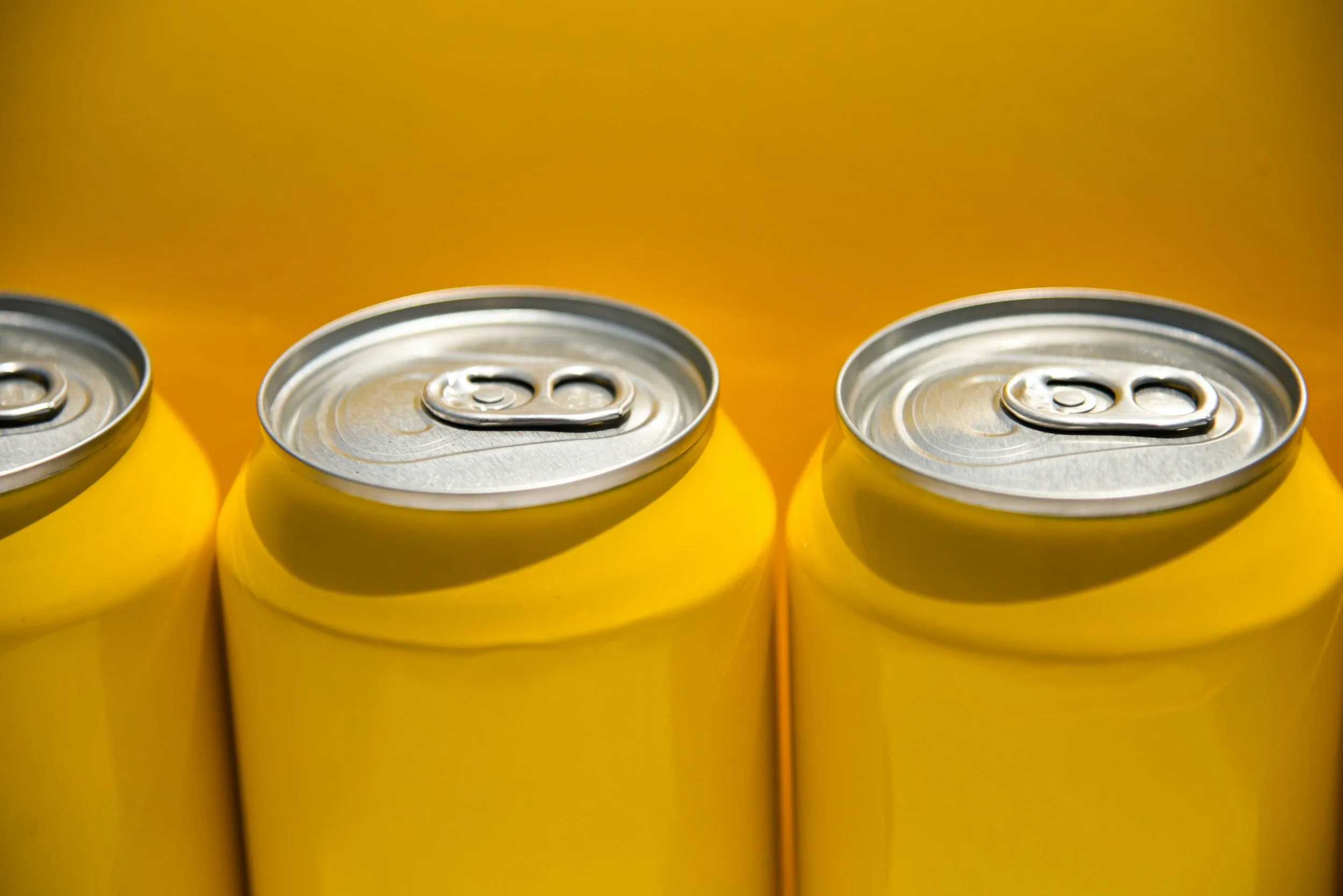 Three yellow soda cans with pull-tab tops, aligned in a row on a yellow background.