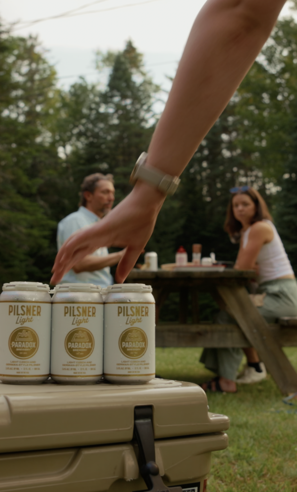 Close-up of four cans of Paradox Pilsner Light beer on a cooler, with a person's hand reaching over them. In the background, two people are sitting at a picnic table outdoors in front of a wooded area, with food and beverages on the table.
