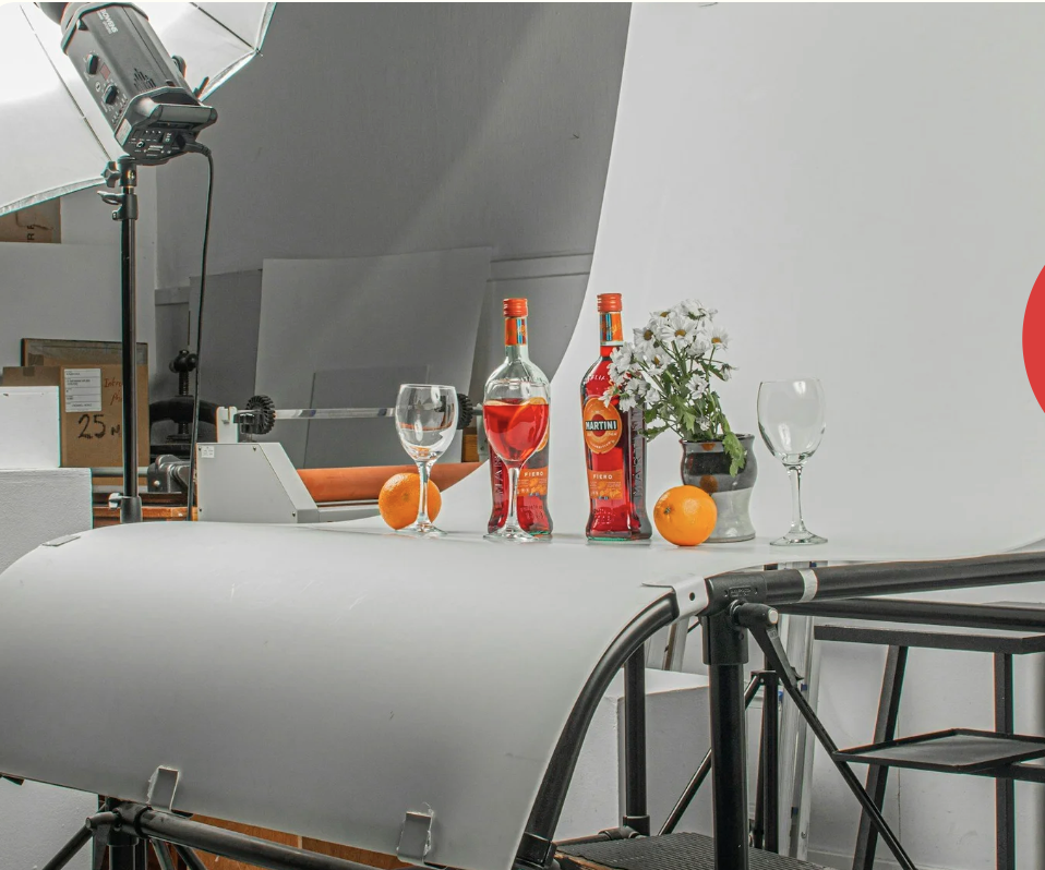 Photography studio setup with a white background, featuring two bottles of martini, two wine glasses, an orange, a vase with white flowers, and a photography light.